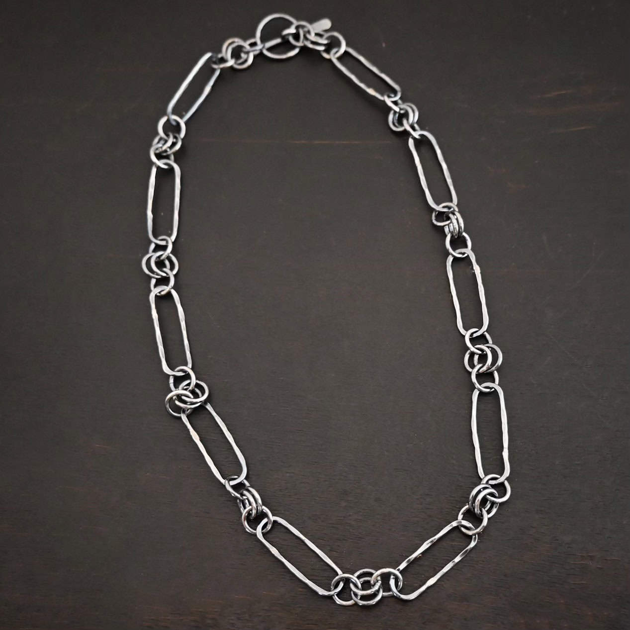 Close up of the "Bekah" Chain. This chain is handmade out of sterling silver and is oxidized. This chain has large oval links that are hammered and connected together by four jump rings. Chain is laying against a dark background.