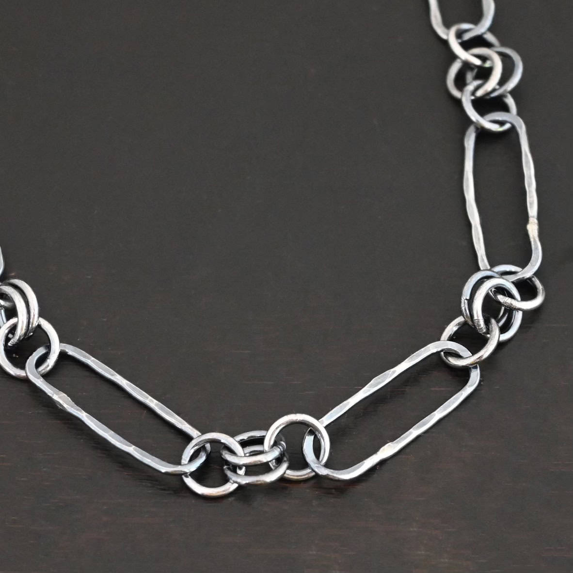 Close up of the "Bekah" Chain. This chain is handmade out of sterling silver and is oxidized. This chain has large oval links that are hammered and connected together by four jump rings. Chain is laying against a dark background.