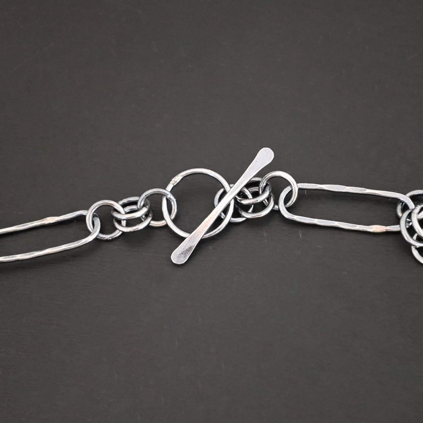Close up of the "Bekah" Chain. This chain is handmade out of sterling silver and is oxidized. This chain has large oval links that are hammered and connected together by four jump rings. Focus is on the toggle clasp that the chain has to connect it. Clasp is laying against a dark background.