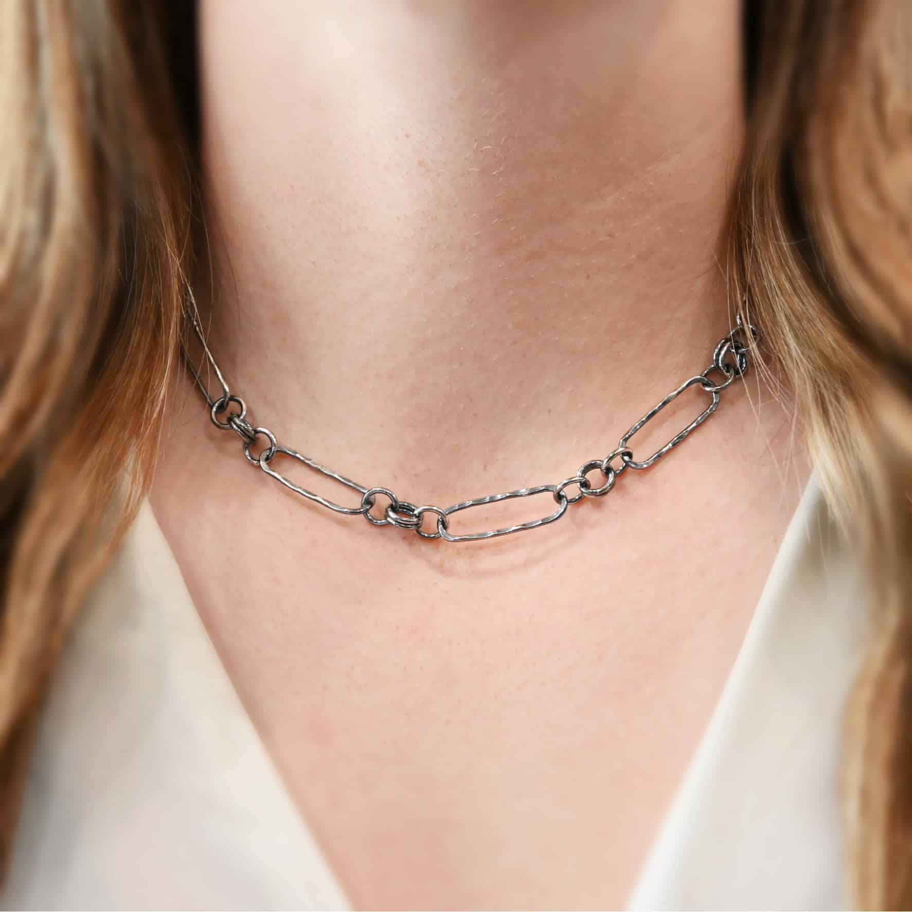 Close up of the "Bekah" Chain as shown on a model. This chain is handmade out of sterling silver and is oxidized. This chain has large oval links that are hammered and connected together by four jump rings. The chain is worn by a model. Chain is 16 inches long. Model is wearing a white v-neck.
