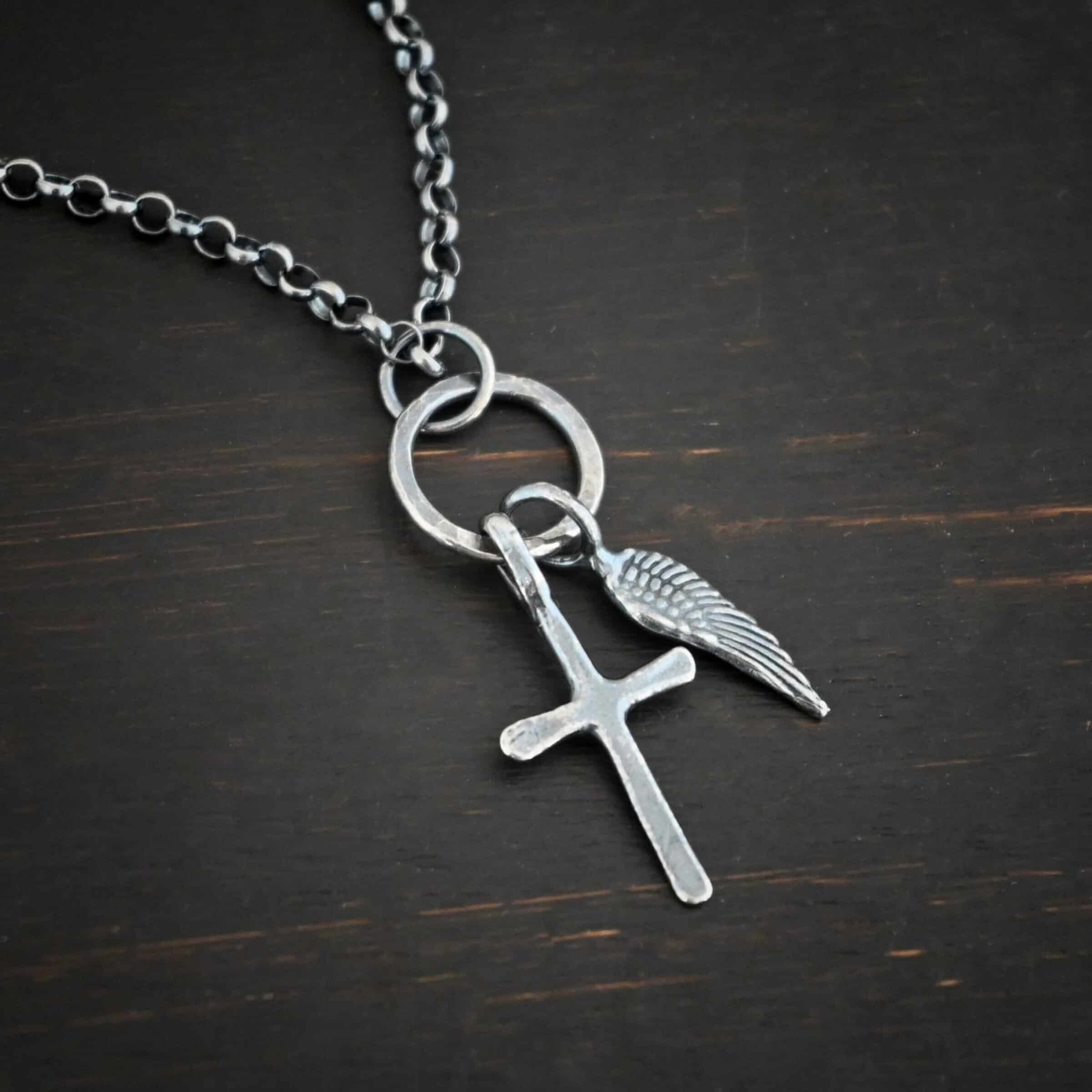 Strength Necklace Photo of the "Strength" Necklace. Made out of sterling silver. There is a small sized cross that is made out of smooth sterling silver. It is oxidized and brushed lightly and has a loop soldered to the top of it. There is a small sized angel wing pendant next to it, also with a soldered loop on the top. The feathers and lines on the angel wing are oxidized to stand out. There is a chain that goes through the top of the necklace. Necklace is lying against a dark background.