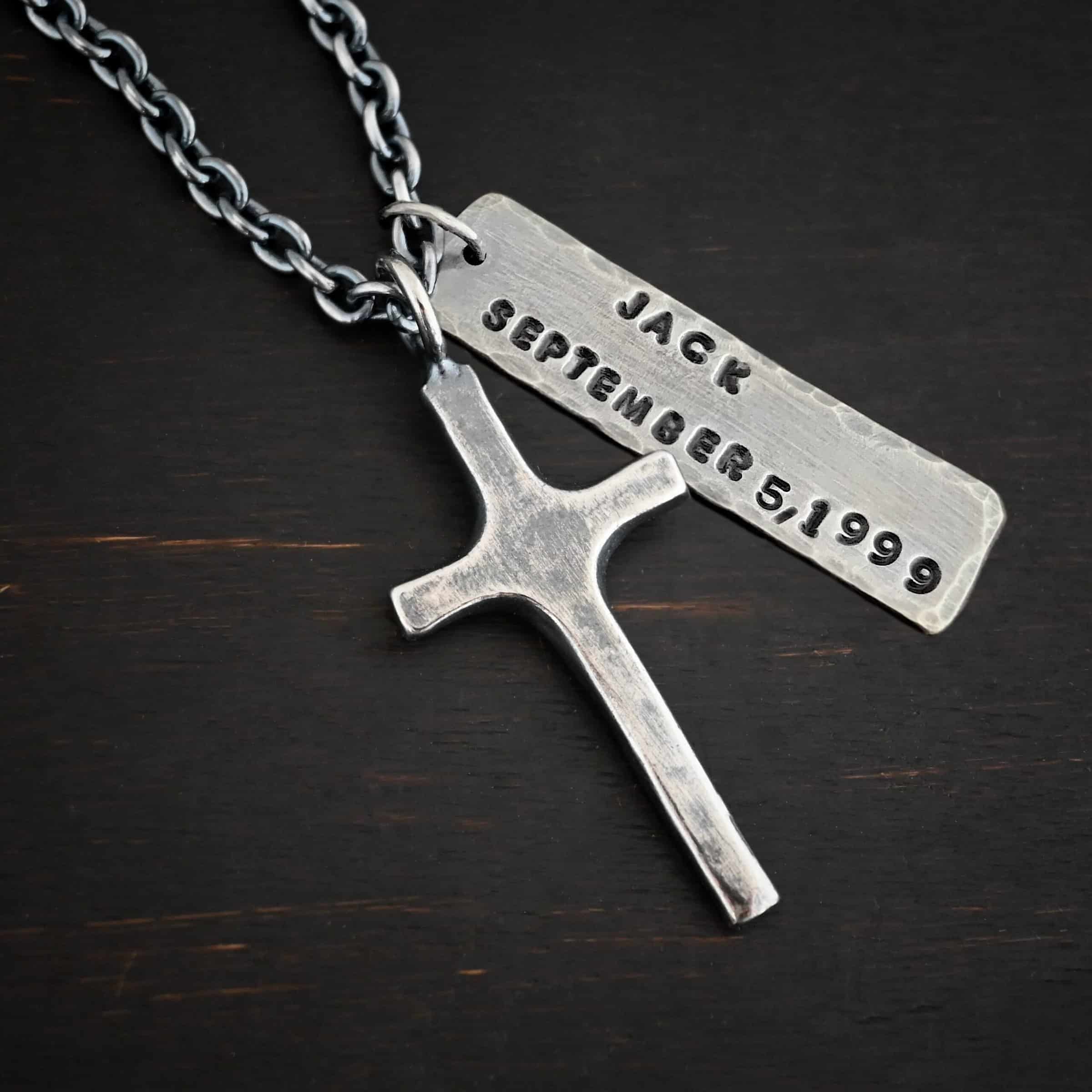 King of Kings Custom Photo of a custom "King Of Kings" Necklace. Made out of sterling silver. There is a medium sized cross that is made out of smooth, thick sterling silver. It is oxidized and brushed lightly and has a loop soldered to the top of it. There is a medium sized piece of rectangular silver next to it. It is oxidized and hammered around the edges. It has the words "Jack, September 5, 1999," stamped into it. The words are darkened. There is a chain that goes through the top of the necklace. Necklace is lying against a dark background.