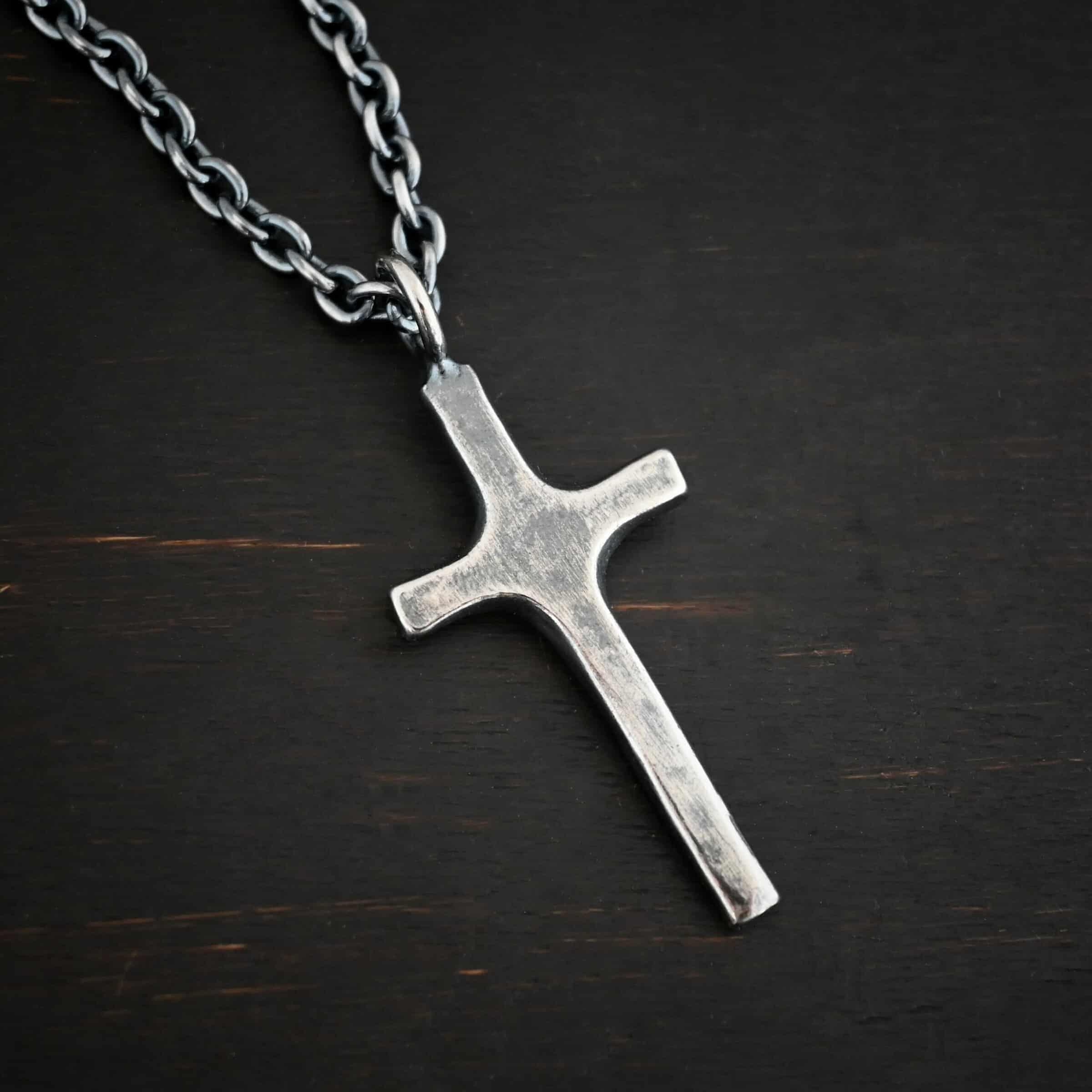 Photo of the "King Of Kings" Necklace. Made out of sterling silver. There is a medium sized cross that is made out of smooth, thick sterling silver. It is oxidized and brushed lightly and has a loop soldered to the top of it. There is a chain that goes through the top of the necklace. Necklace is lying against a dark background.