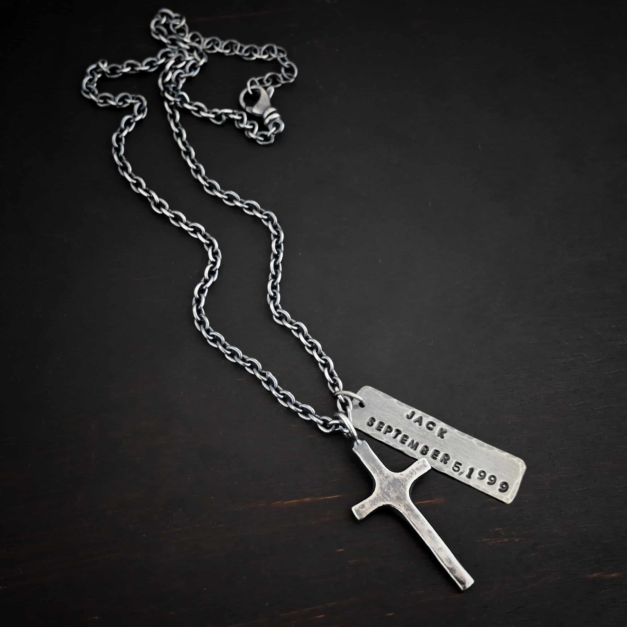 King of Kings Custom Photo of a custom "King Of Kings" Necklace. Made out of sterling silver. There is a medium sized cross that is made out of smooth, thick sterling silver. It is oxidized and brushed lightly and has a loop soldered to the top of it. There is a medium sized piece of rectangular silver next to it. It is oxidized and hammered around the edges. It has the words "Jack, September 5, 1999," stamped into it. The words are darkened. There is a chain that goes through the top of the necklace. Necklace is lying against a dark background.