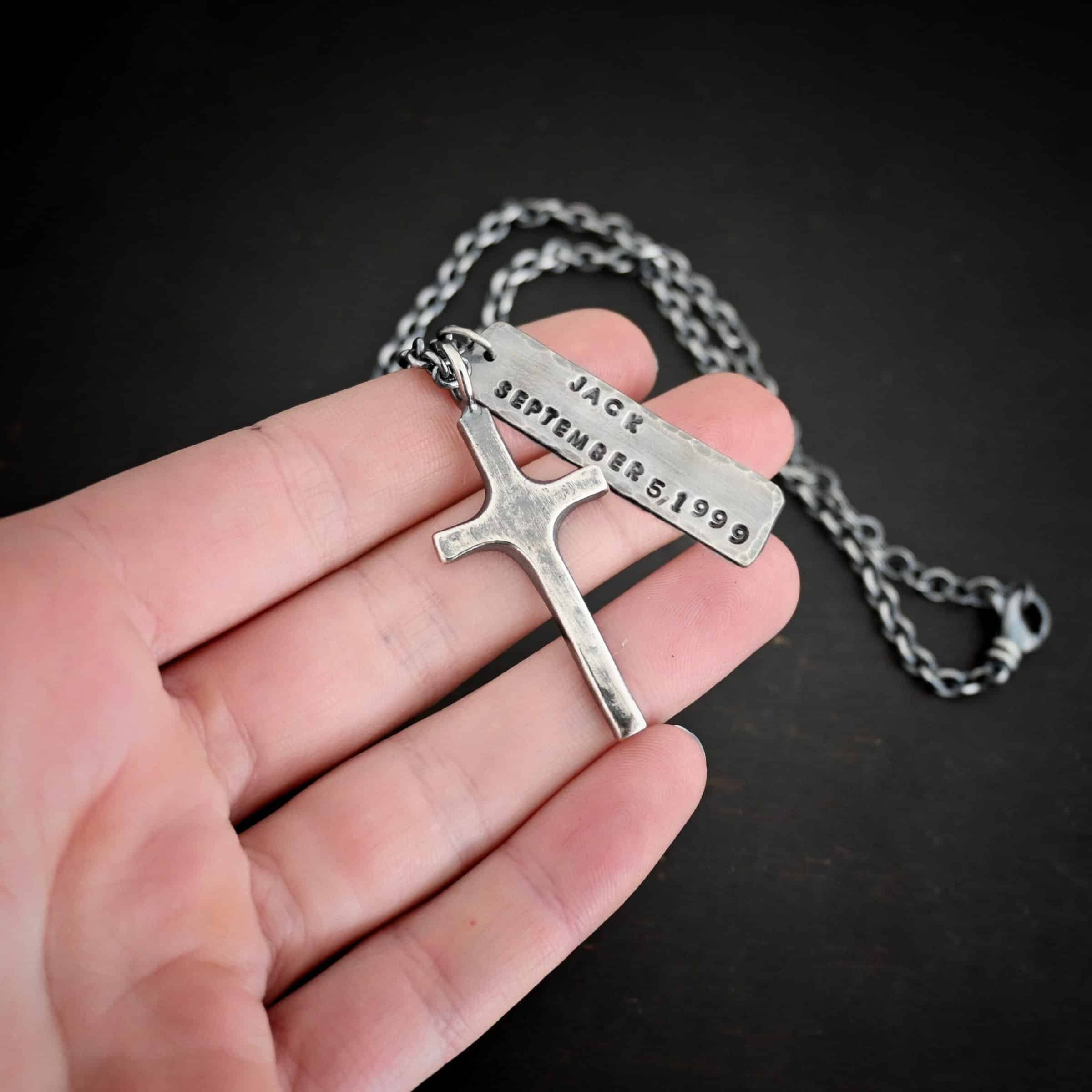 King of Kings Custom Photo of a custom "King Of Kings" Necklace. Made out of sterling silver. There is a medium sized cross that is made out of smooth, thick sterling silver. It is oxidized and brushed lightly and has a loop soldered to the top of it. There is a medium sized piece of rectangular silver next to it. It is oxidized and hammered around the edges. It has the words "Jack, September 5, 1999," stamped into it. The words are darkened. There is a chain that goes through the top of the necklace. Necklace is lying on the fingertips of a hand to demonstrate size.
