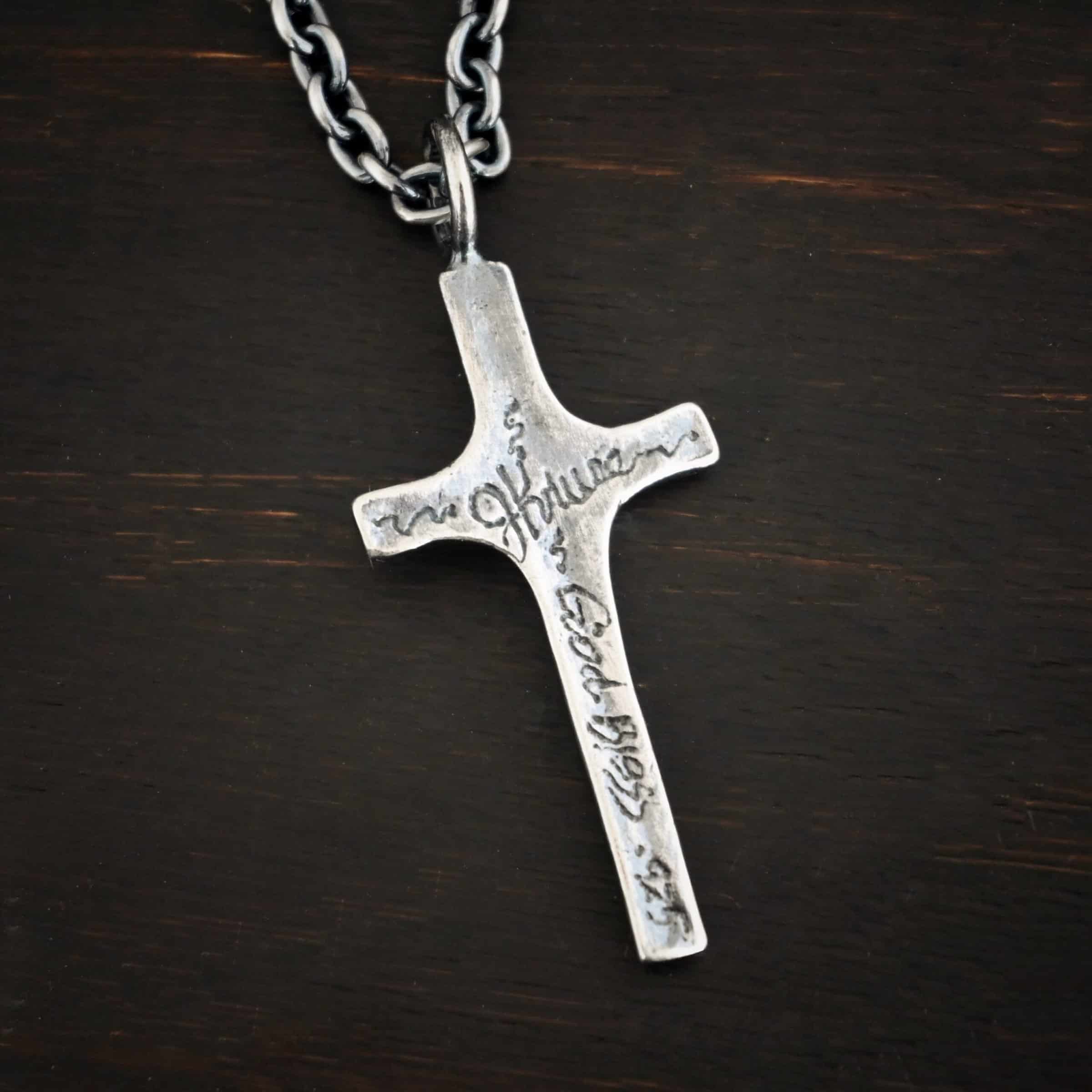 Photo of the back of the"King Of Kings" Necklace. Made out of sterling silver. There is a medium sized cross that is made out of smooth, thick sterling silver. It is oxidized and brushed lightly and has a loop soldered to the top of it. The words "JKruse - God Bless - .925" engraved into it. There is a chain that goes through the top of the necklace. Necklace is lying against a dark background.