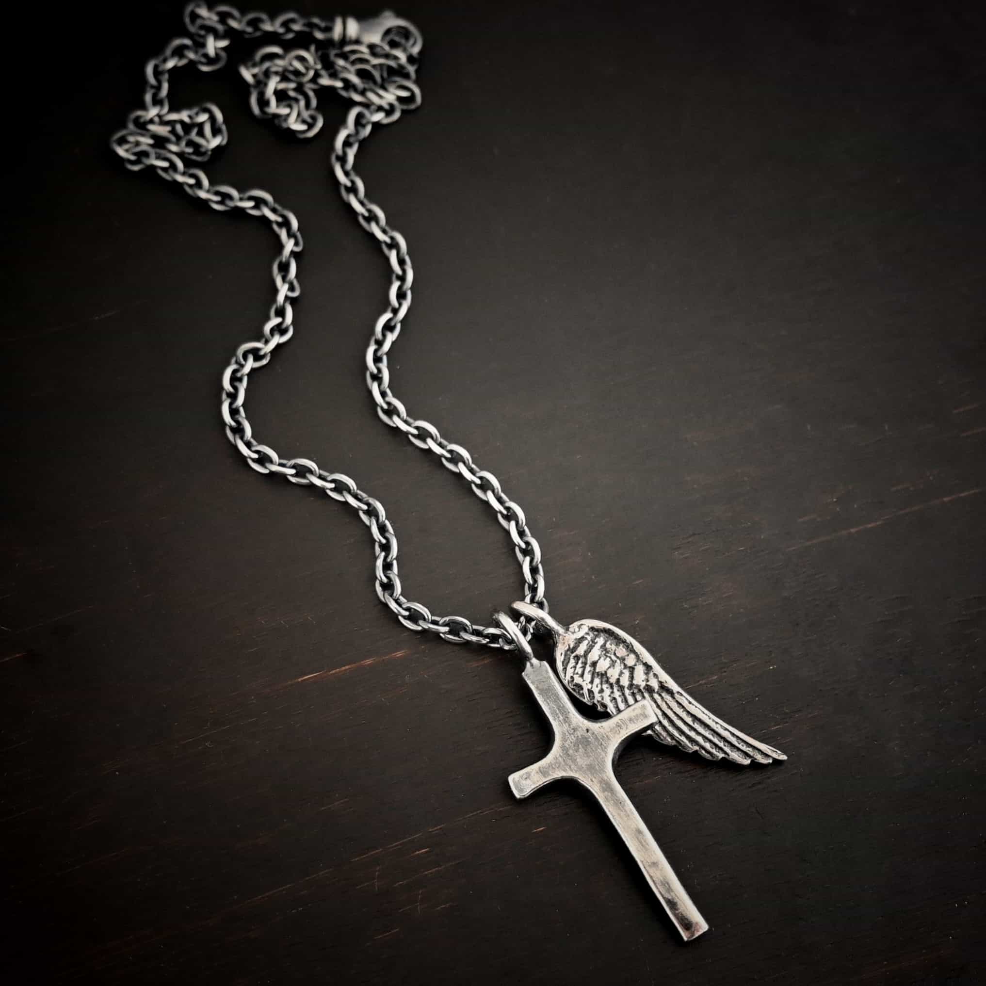Photo of the "Guardian Angel" Necklace. Made out of sterling silver. There is a medium sized cross that is made out of smooth, thick sterling silver. It is oxidized and brushed lightly and has a loop soldered to the top of it. There is a medium sized angel wing pendant next to it, also with a soldered loop on the top. The feathers and lines on the angel wing are oxidized to stand out. There is a chain that goes through the top of the necklace. Necklace is lying against a dark background.