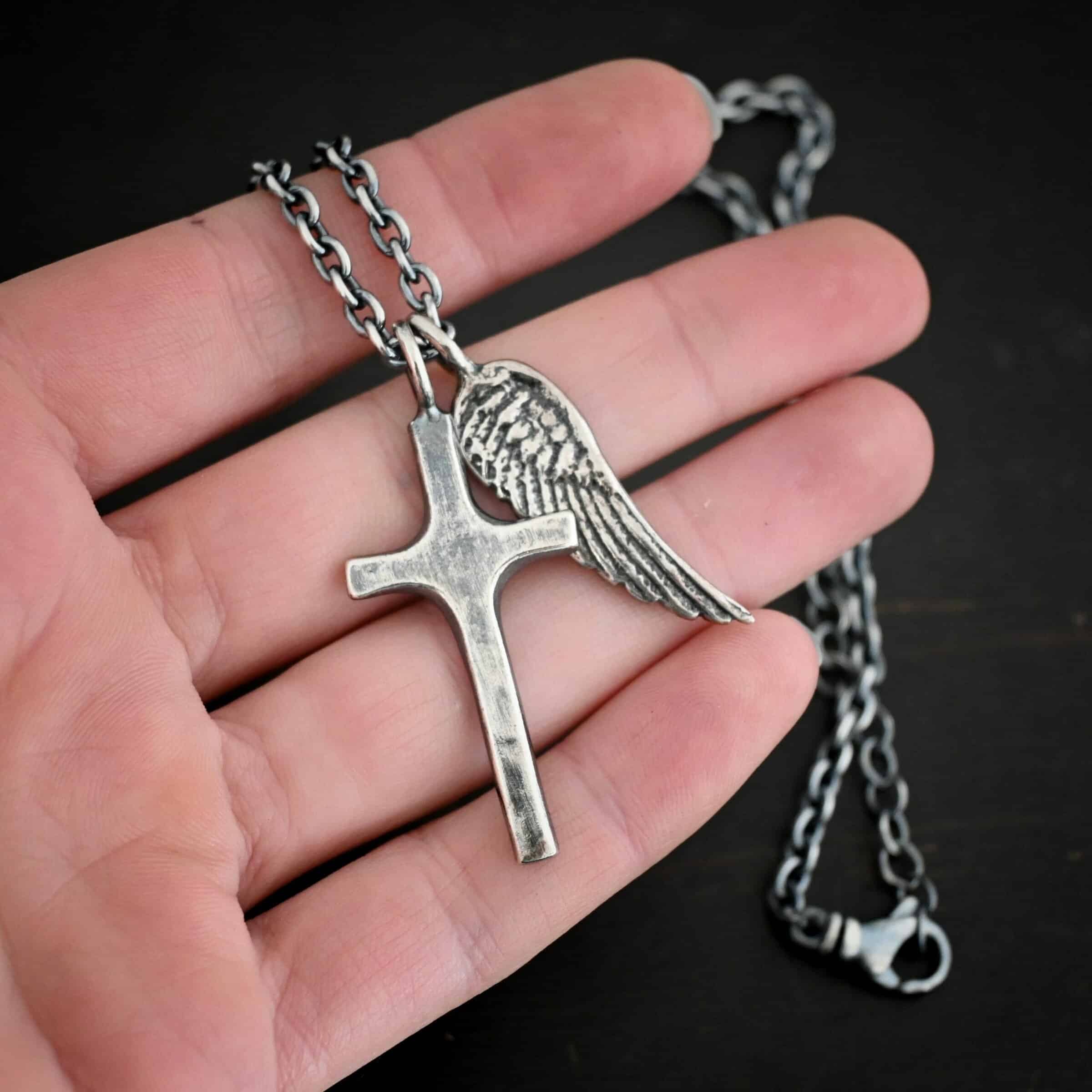 Photo of the "Guardian Angel" Necklace. Made out of sterling silver. There is a medium sized cross that is made out of smooth, thick sterling silver. It is oxidized and brushed lightly and has a loop soldered to the top of it. There is a medium sized angel wing pendant next to it, also with a soldered loop on the top. The feathers and lines on the angel wing are oxidized to stand out. There is a chain that goes through the top of the necklace. Necklace is lying against the fingertips of a hand.