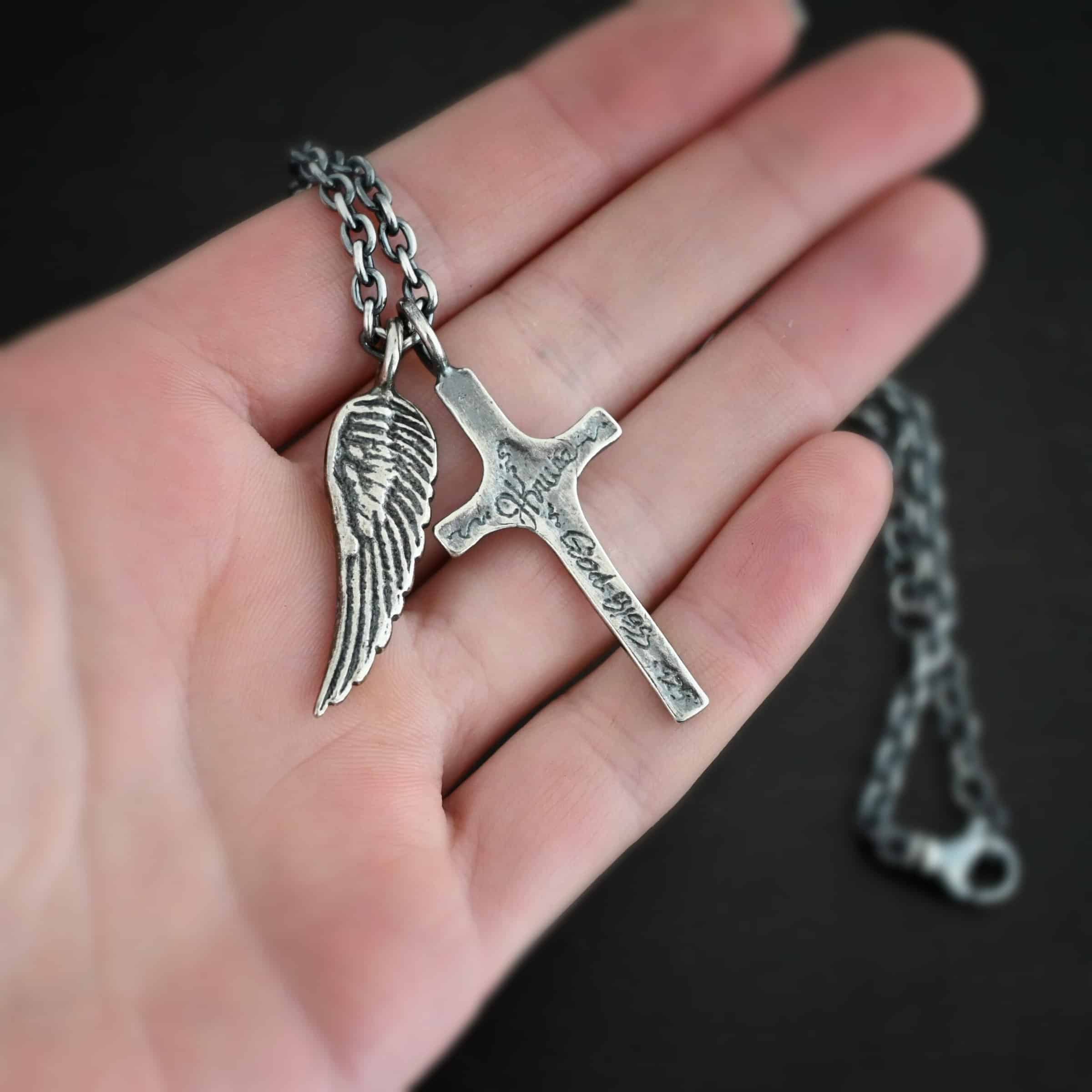 Photo of the "Guardian Angel" Necklace. Made out of sterling silver. There is a medium sized cross that is made out of smooth, thick sterling silver with the name "JKruse" and "God Bless" and ".925" engraved into it. It is oxidized and brushed lightly and has a loop soldered to the top of it. There is a medium sized angel wing pendant next to it, also with a soldered loop on the top. The feathers and lines on the angel wing are oxidized to stand out. There is a chain that goes through the top of the necklace. Necklace is lying against the fingertips of a hand.
