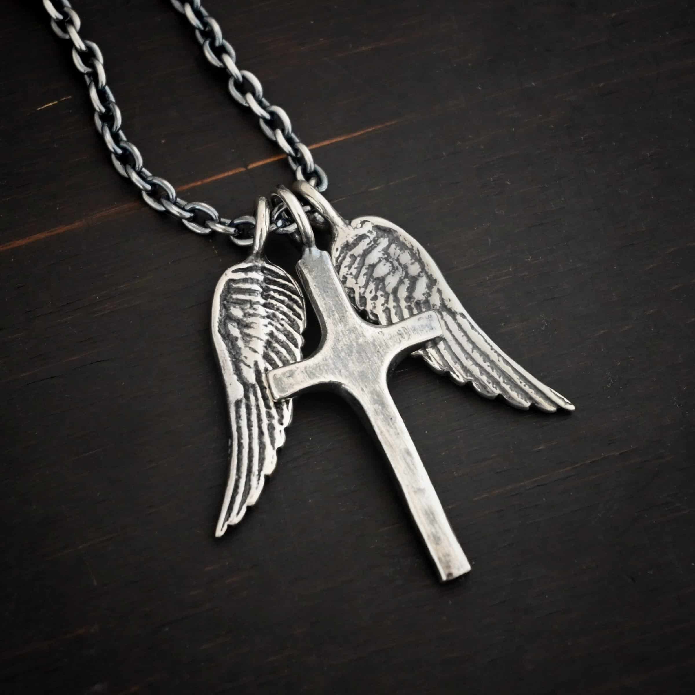 Photo of the "Guardian Angel" Necklace. Made out of sterling silver. There is a medium sized cross that is made out of smooth, thick sterling silver. It is oxidized and brushed lightly and has a loop soldered to the top of it. There are two medium sized angel wing pendants next to it, also with soldered loops on the top. The feathers and lines on the angel wing are oxidized to stand out. Each wing is on a different side of the cross. There is a chain that goes through the top of the necklace. Necklace is lying against a dark background.
