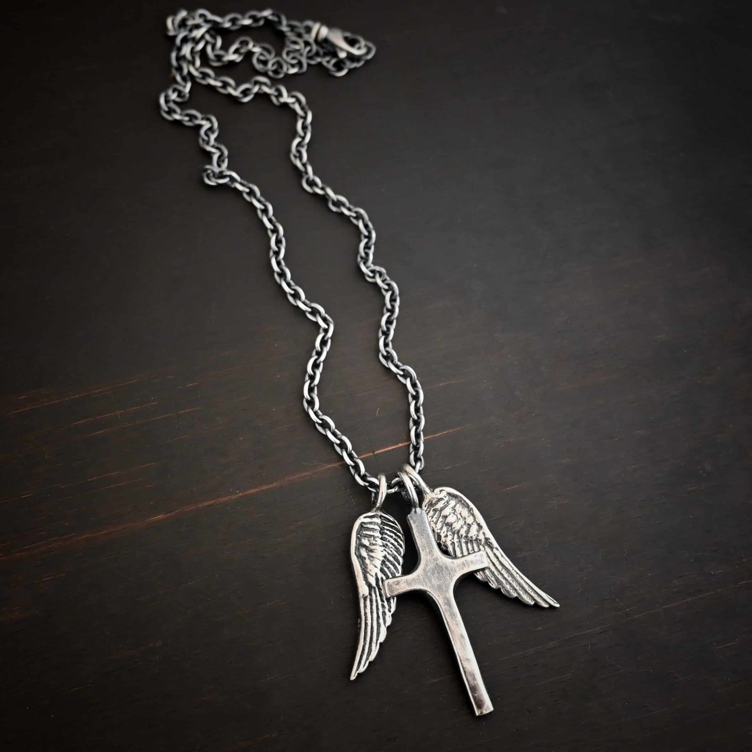 Photo of the "Guardian Angel" Necklace. Made out of sterling silver. There is a medium sized cross that is made out of smooth, thick sterling silver. It is oxidized and brushed lightly and has a loop soldered to the top of it. There are two medium sized angel wing pendants next to it, also with soldered loops on the top. The feathers and lines on the angel wing are oxidized to stand out. Each wing is on a different side of the cross. There is a chain that goes through the top of the necklace. Necklace is lying against a dark background.