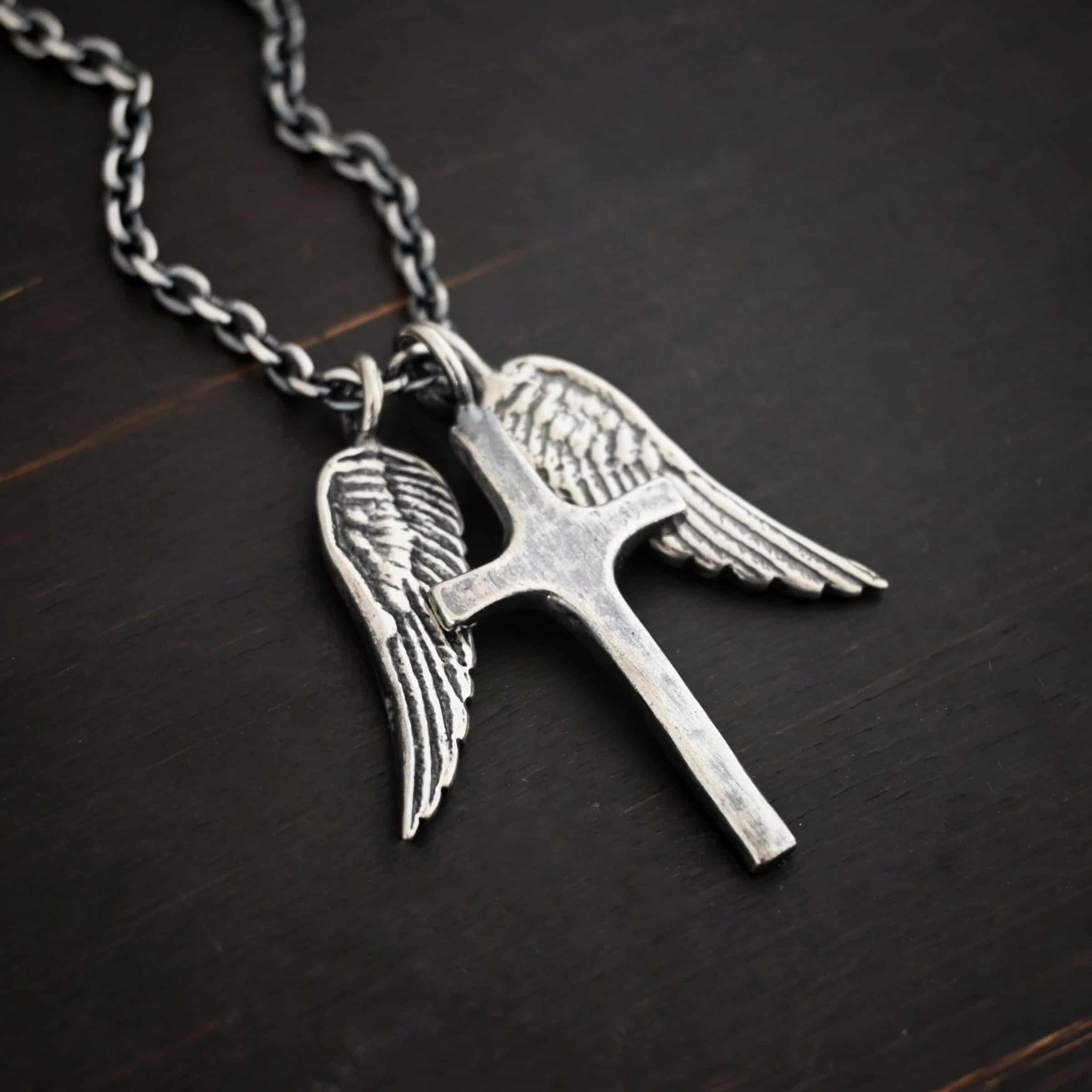 Photo of the "Guardian Angel" Necklace. Made out of sterling silver. There is a medium sized cross that is made out of smooth, thick sterling silver. It is oxidized and brushed lightly and has a loop soldered to the top of it. There are two medium sized angel wing pendants next to it, also with soldered loops on the top. The feathers and lines on the angel wing are oxidized to stand out. Each wing is on a different side of the cross. There is a chain that goes through the top of the necklace. Necklace is lying against a dark background.