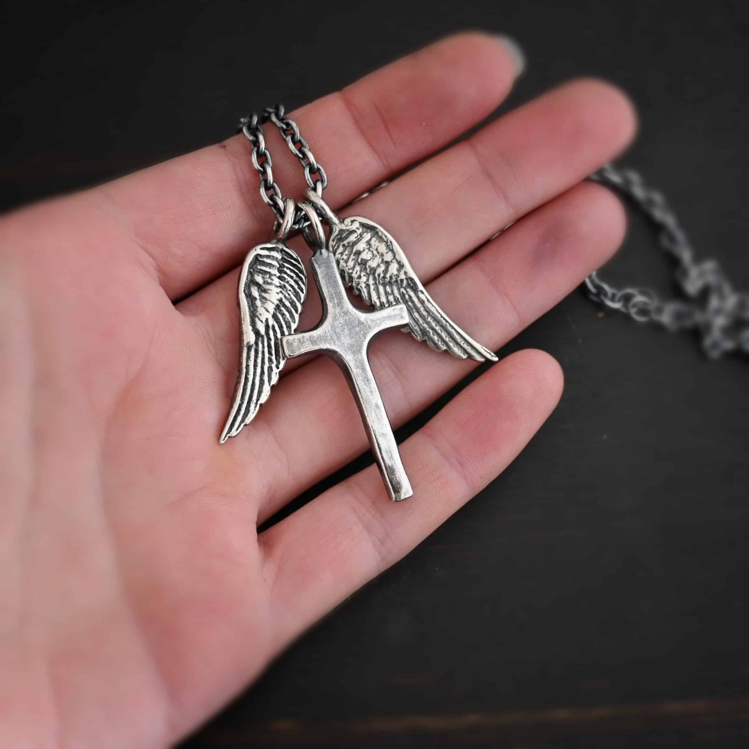 Photo of the "Guardian Angel" Necklace. Made out of sterling silver. There is a medium sized cross that is made out of smooth, thick sterling silver. It is oxidized and brushed lightly and has a loop soldered to the top of it. There are two medium sized angel wing pendants next to it, also with soldered loops on the top. The feathers and lines on the angel wing are oxidized to stand out. Each wing is on a different side of the cross. There is a chain that goes through the top of the necklace. Necklace is lying against the fingertips of a hand.