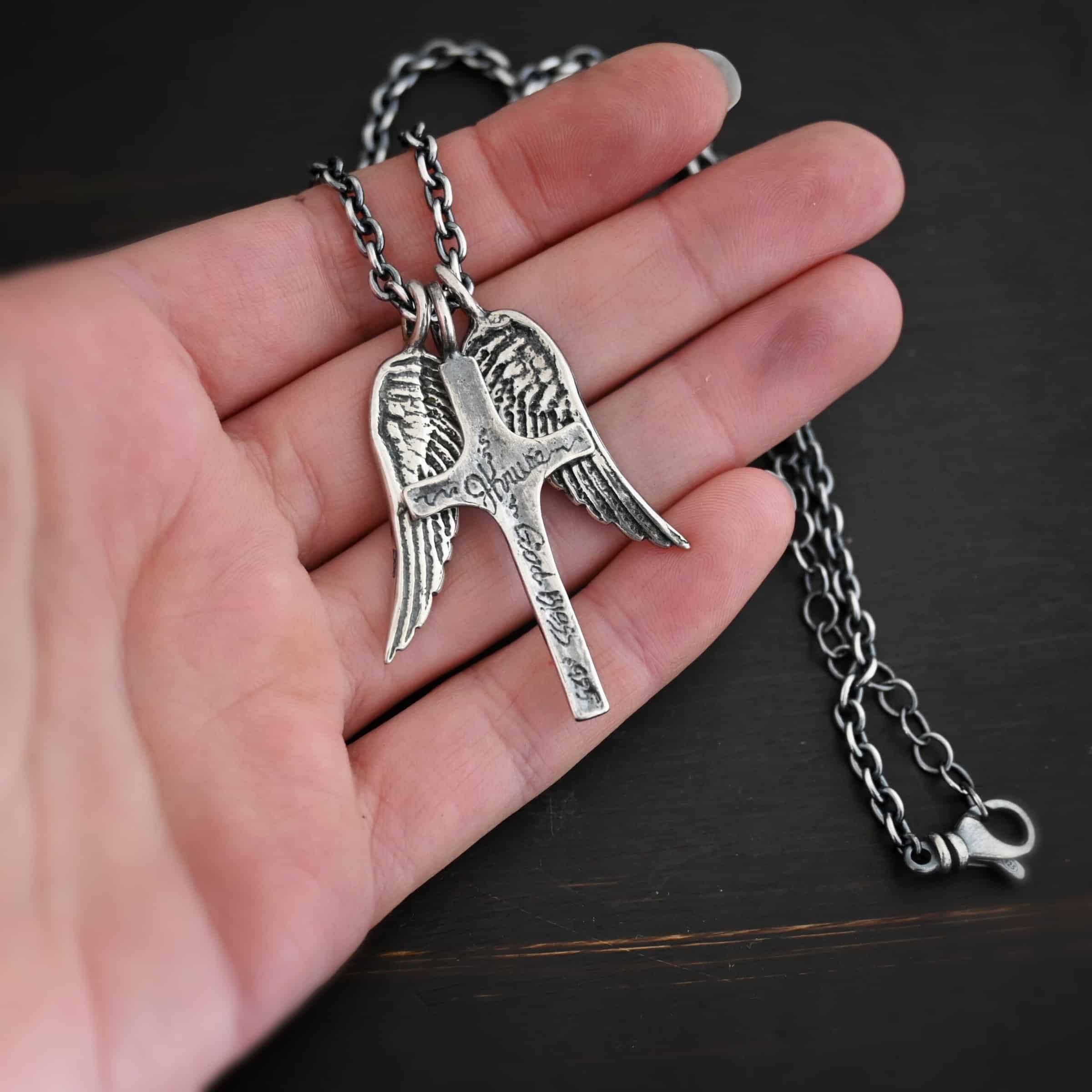 Photo of the "Guardian Angel" Necklace. Made out of sterling silver. There is a medium sized cross that is made out of smooth, thick sterling silver. It is oxidized and brushed lightly and has a loop soldered to the top of it. It has the name "JKruse" engraved into it, as well as the words "God Bless" and ".925." There are two medium sized angel wing pendants next to it, also with soldered loops on the top. The feathers and lines on the angel wing are oxidized to stand out. Each wing is on a different side of the cross. There is a chain that goes through the top of the necklace. Necklace is lying against the fingertips of a hand.