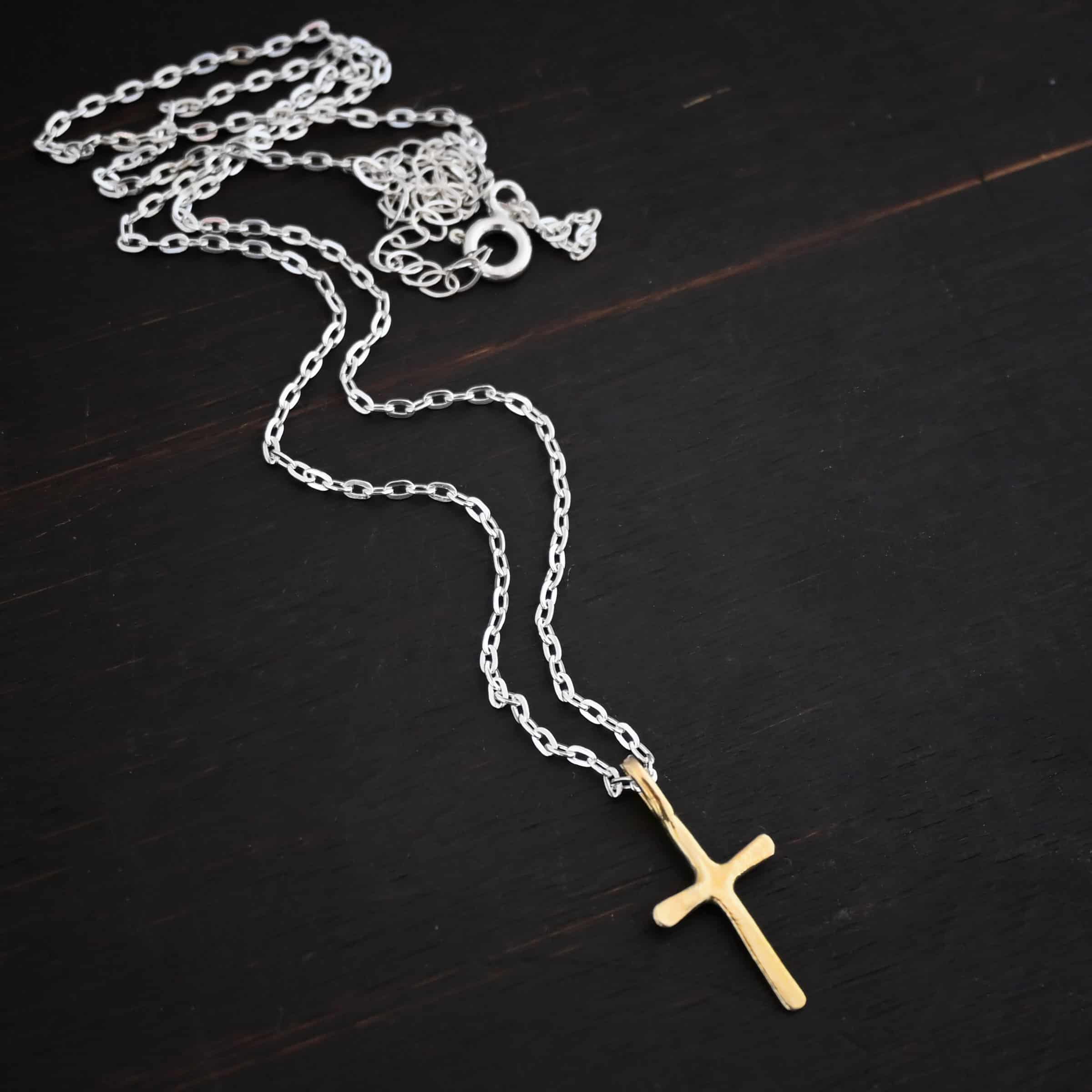 gold shiny redeemer Photo of the "Redeemer" Necklace. Made out of 18k gold. Necklace has a very small cross that is hammered flat, but smooth, with a loop soldered to the top of it. The chain of the necklace is shiny and has a spring ring clasp. Necklace is lying against a dark background.