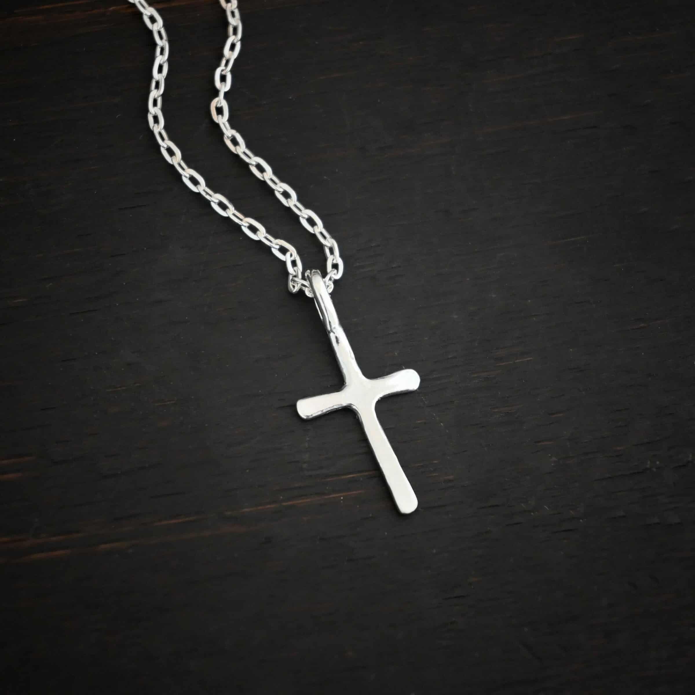 silver shiny redeemer Photo of the "Redeemer" Necklace. Made out of sterling silver and shiny. Necklace has a very small cross that is hammered flat, but smooth, with a loop soldered to the top of it. The chain of the necklace is shiny. Necklace is lying against a dark background.