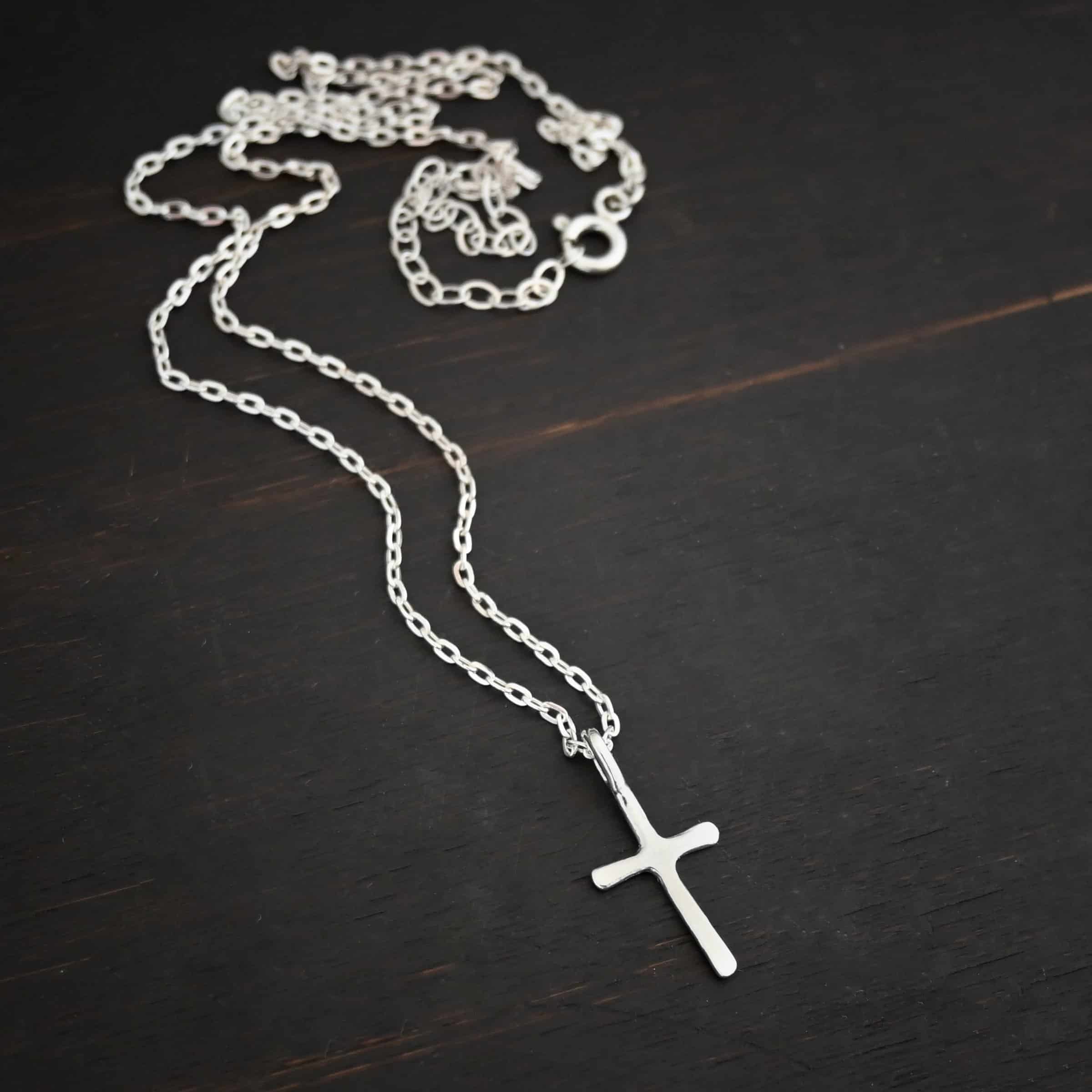silver shiny redeemer Photo of the "Redeemer" Necklace. Made out of sterling silver and shiny. Necklace has a very small cross that is hammered flat, but smooth, with a loop soldered to the top of it. The chain of the necklace is shiny and has a spring ring clasp. Necklace is lying against a dark background.
