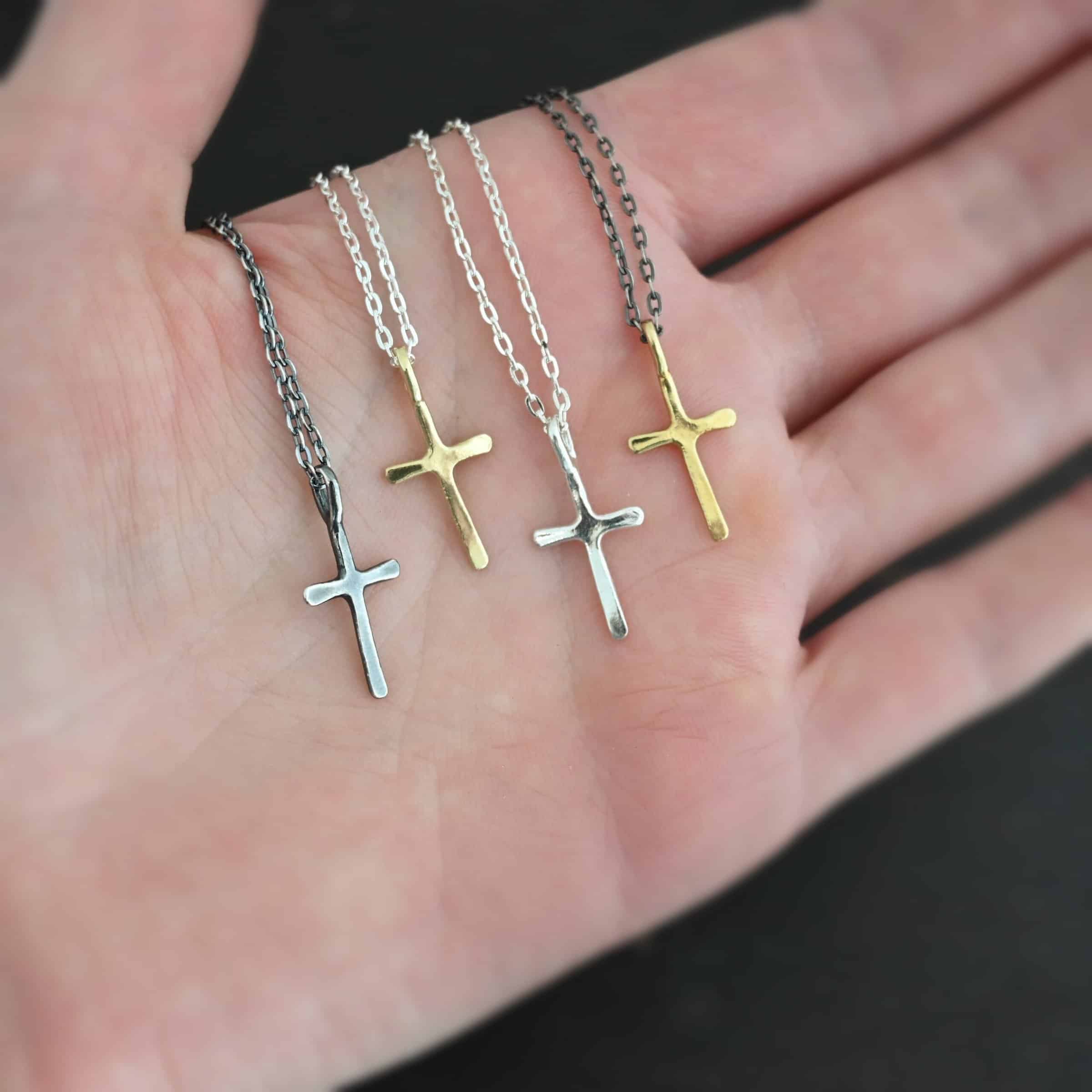 Redeemer Necklace Photo of four of the "Redeemer" Necklaces. Made out of sterling silver and gold. Each necklace has a very small cross that is hammered flat, but smooth, with a loop soldered to the top of it. The first cross is oxidized, the second is gold, the third is shiny, and the final is gold again. The first chain of the necklace is oxidized, the second two are shiny, and the final is oxidized. Each necklace is lying against the palm of a hand.