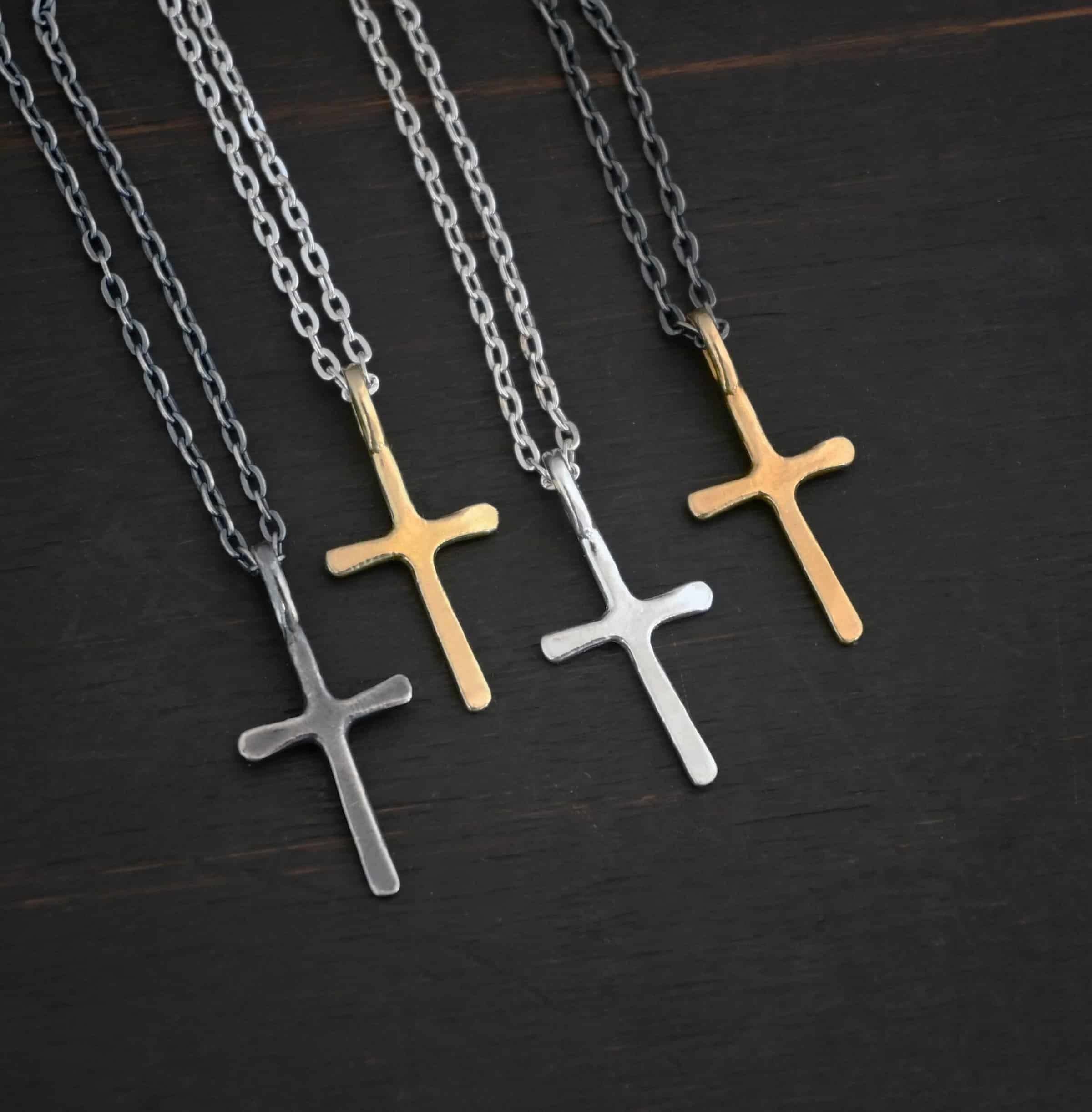 Redeemer Necklace Photo of four of the "Redeemer" Necklaces. Made out of sterling silver and gold. Each necklace has a very small cross that is hammered flat, but smooth, with a loop soldered to the top of it. The first cross is oxidized, the second is gold, the third is shiny, and the final is gold again. The first chain of the necklace is oxidized, the second two are shiny, and the final is oxidized. Each necklace is lying against a dark background.