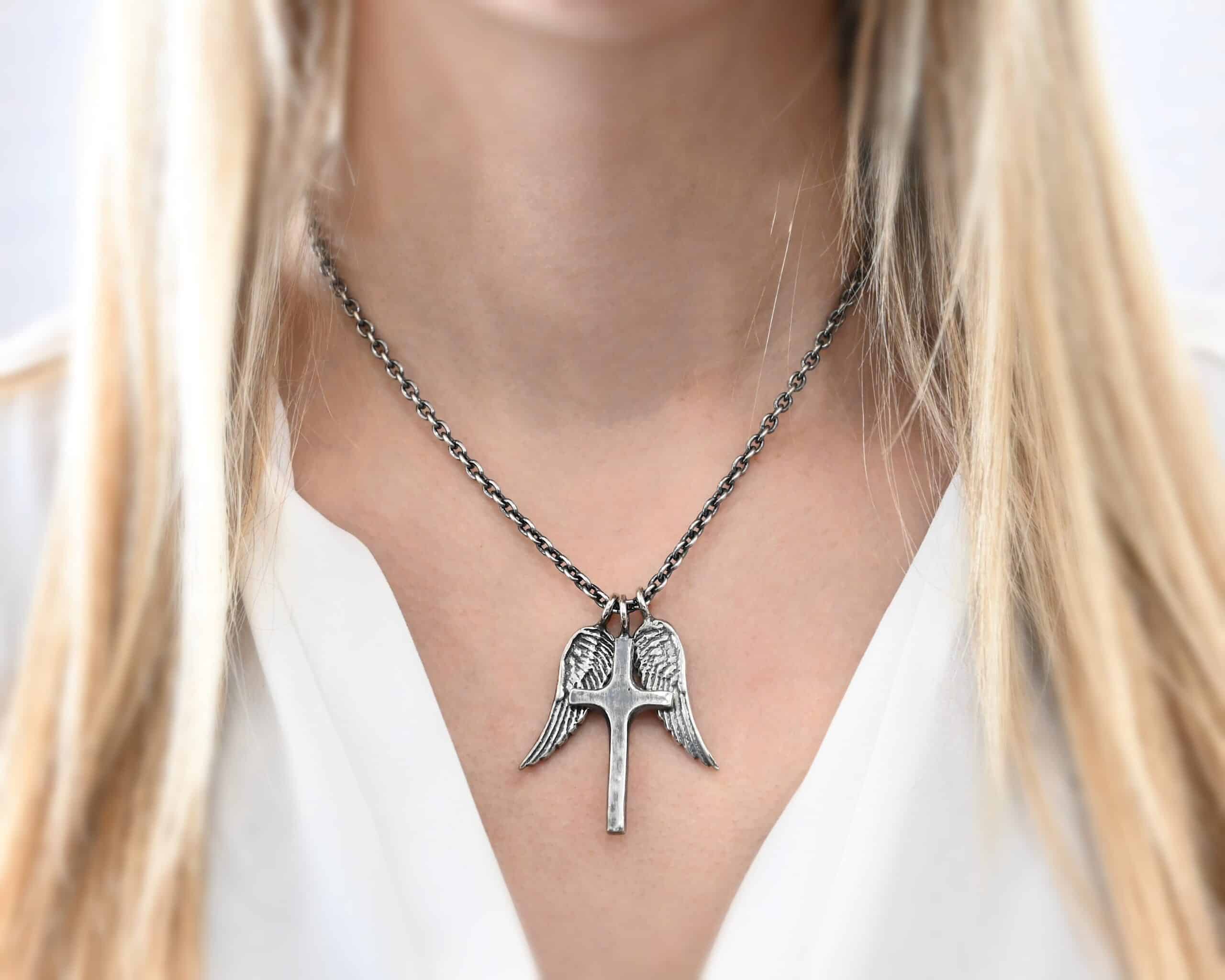 Photo of the "Guardian Angel" Necklace. Made out of sterling silver. There is a medium sized cross that is made out of smooth, thick sterling silver. It is oxidized and brushed lightly and has a loop soldered to the top of it. There are two medium sized angel wing pendants next to it, also with soldered loops on the top. The feathers and lines on the angel wing are oxidized to stand out. Each wing is on a different side of the cross. There is a chain that goes through the top of the necklace. Necklace is worn by a model.