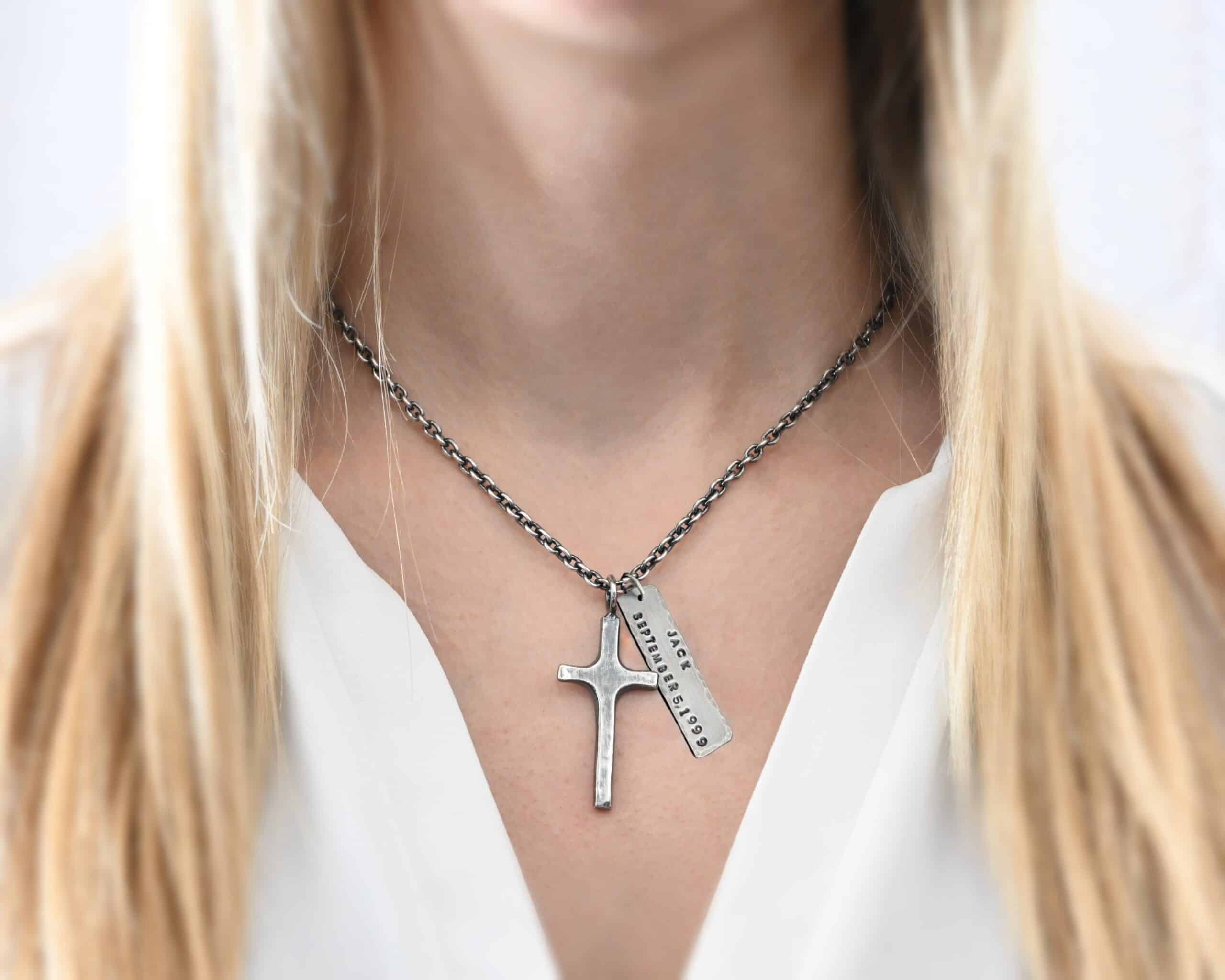 King of Kings Custom Photo of a custom "King Of Kings" Necklace. Made out of sterling silver. There is a medium sized cross that is made out of smooth, thick sterling silver. It is oxidized and brushed lightly and has a loop soldered to the top of it. There is a medium sized piece of rectangular silver next to it. It is oxidized and hammered around the edges. It has the words "Jack, September 5, 1999," stamped into it. The words are darkened. There is a chain that goes through the top of the necklace. Necklace is worn by a model.