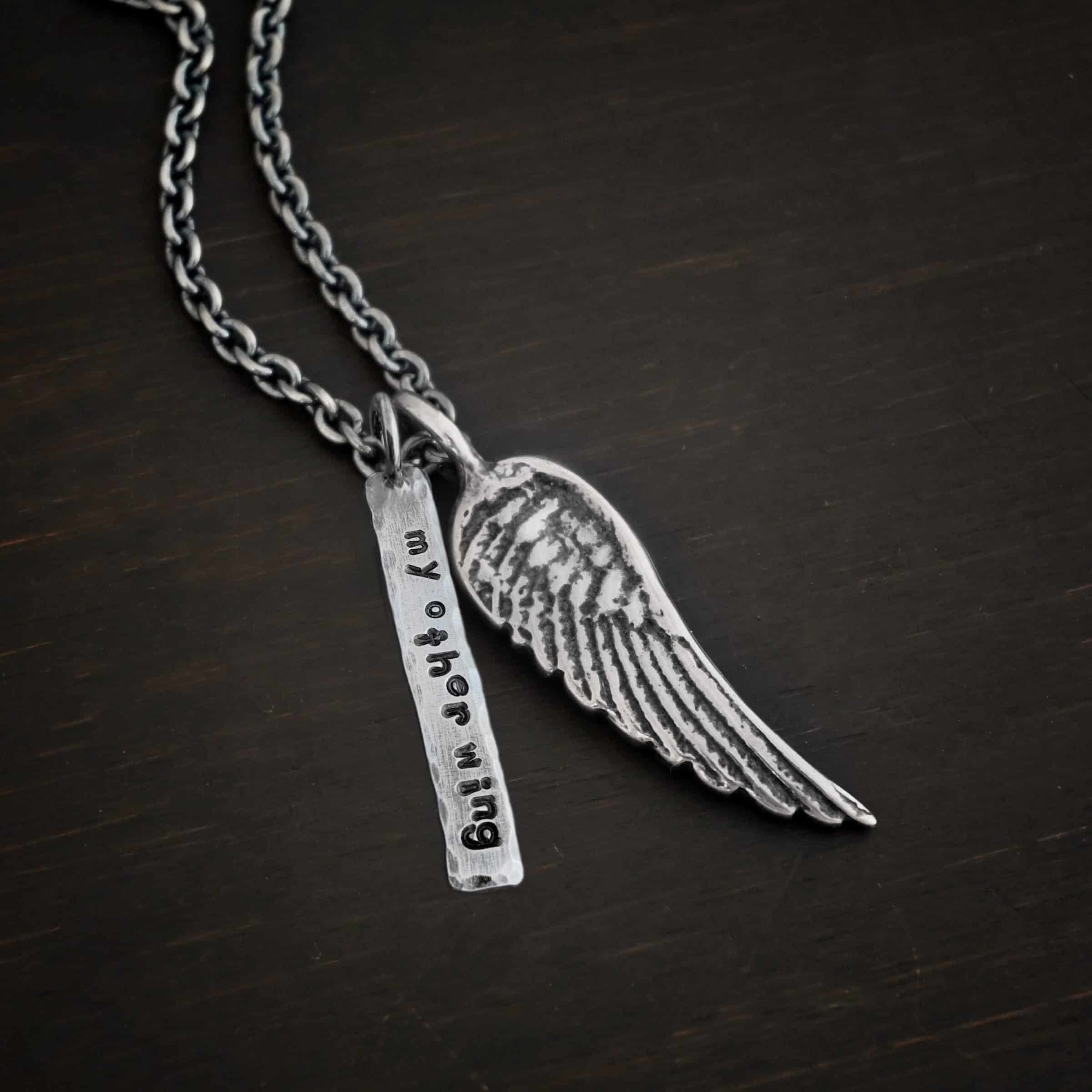 Angel Kiss Necklace with tag Photo of the "Angel Kiss" Necklace. Made out of sterling silver. There is a medium sized angel wing pendant with a loop soldered to the top. There is a medium sized rectangle pendant next to it, also with a loop on the top. The rectangle's edges are hammered and the entire rectangle is oxidized. It has the words "My Other Wing" stamped into it and darkened. There is a chain going through both of the charms. Necklace is lying against a dark background.