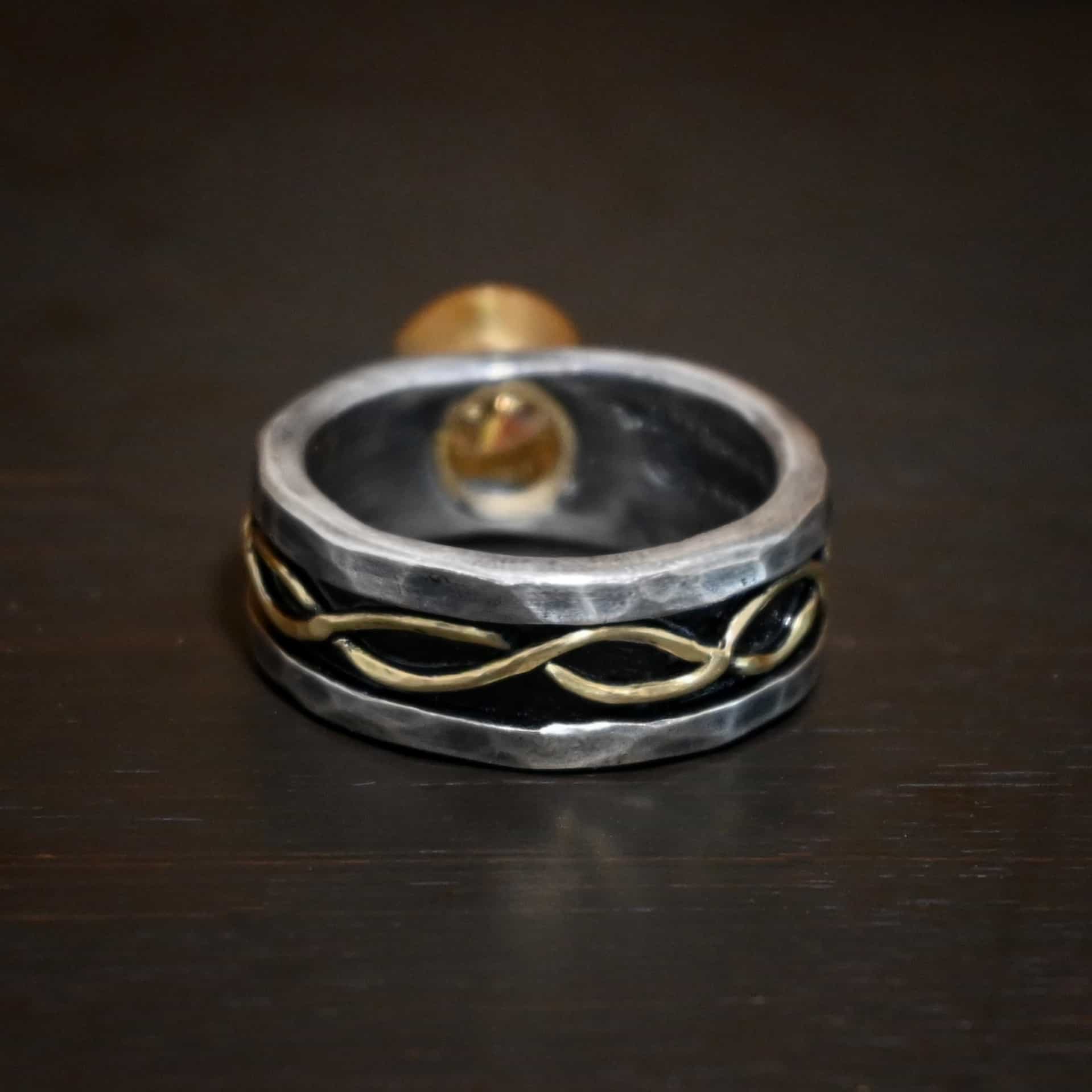 Intertwined Wedding Band Photo of the back of the "Intertwined" wedding band. Each side of the ring has two gold lines that intertwine around each other and meet at the back of the ring. The lines start on the left side, and continues across the back to the right side. The top and bottom of the ring are hammered and lightly oxidized sterling silver. The center of the ring is deeper, and oxidized darker. This is where the gold starts and goes around the entire ring through the center. Ring is sitting against a dark background.