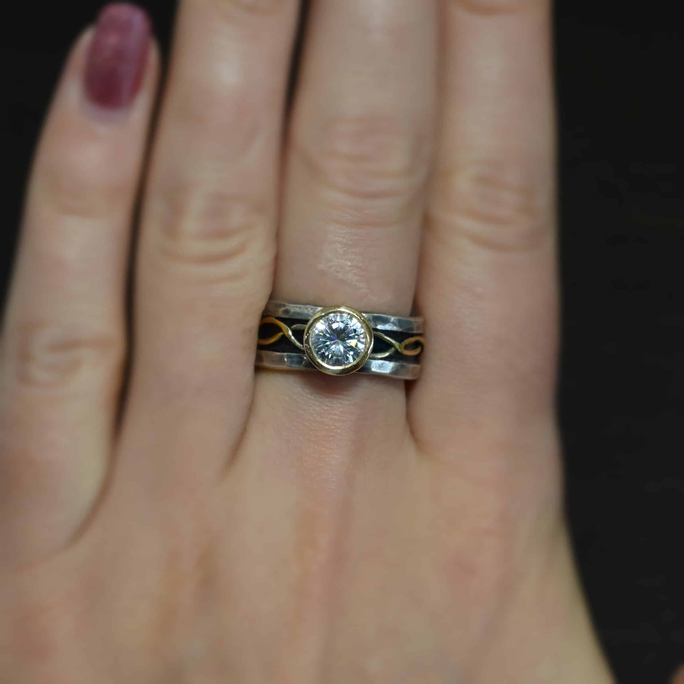 Intertwined Wedding Band Photo of an "Intertwined" wedding band. There is a moissanite that is set in gold in the center of the ring. Each side of the ring has two gold lines that intertwine around each other and meet at the back of the ring. The top and bottom of the ring are hammered and lightly oxidized sterling silver. The center of the ring is deeper, and oxidized darker. This is where the gold starts and goes around the entire ring through the center. Ring is worn by a model.