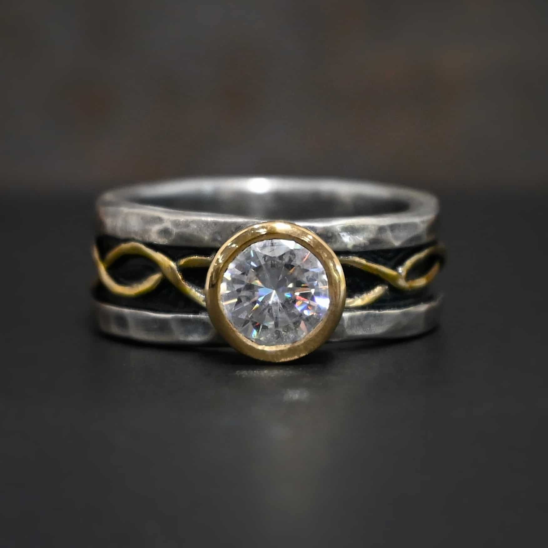 Intertwined Wedding Band Photo of an "Intertwined" wedding band. There is a moissanite that is set in gold in the center of the ring. Each side of the ring has two gold lines that intertwine around each other and meet at the back of the ring. The top and bottom of the ring are hammered and lightly oxidized sterling silver. The center of the ring is deeper, and oxidized darker. This is where the gold starts and goes around the entire ring through the center. Ring is sitting against a dark background.