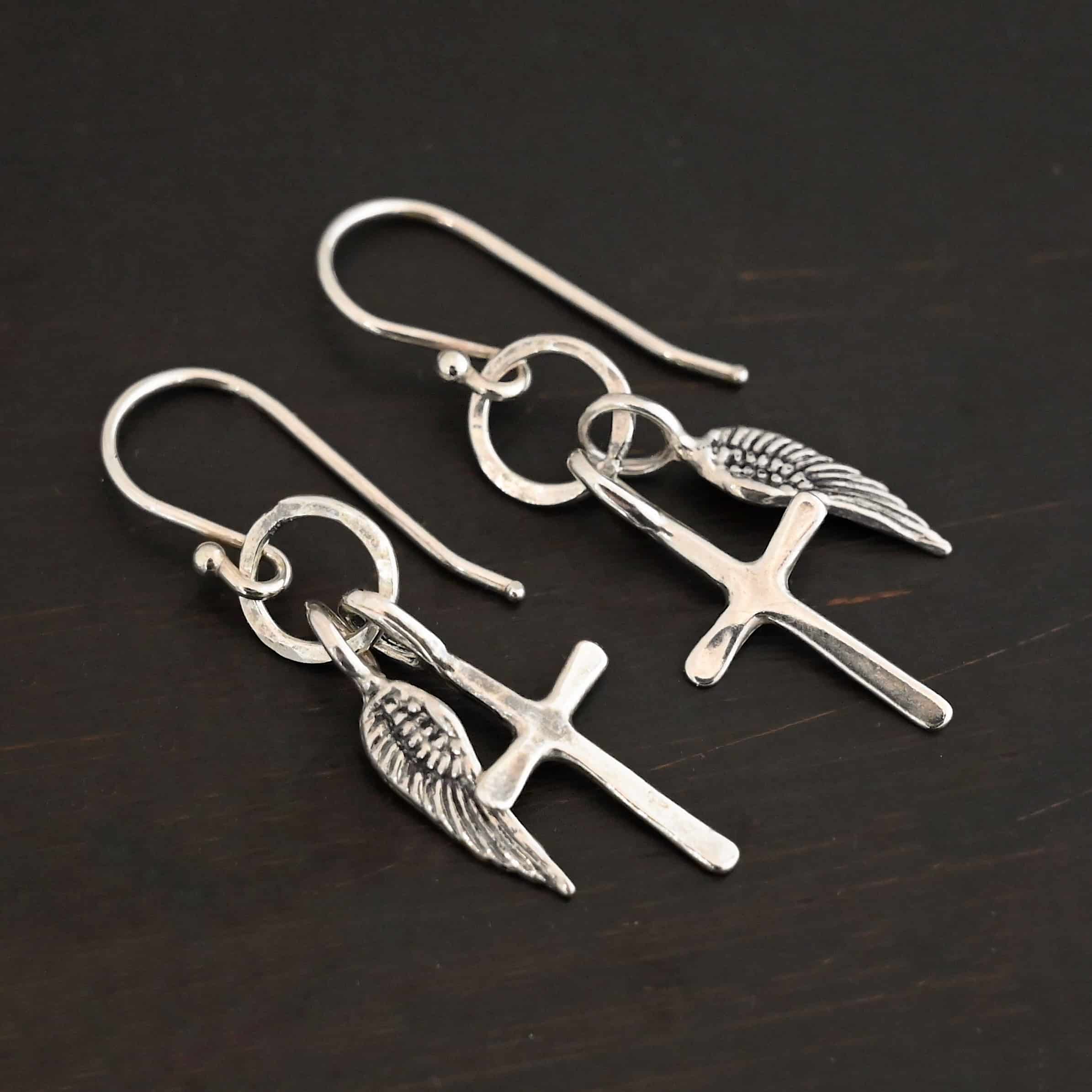 Strength Earrings Photo of the "Strength" Earrings. Made out of sterling silver. There are a cross and angel wing charm on each earring. Both charms are small, and attached to a small hammered sterling silver loop that the French hook ear wires are attached to on the top. Earrings are shiny and laying against a dark background.