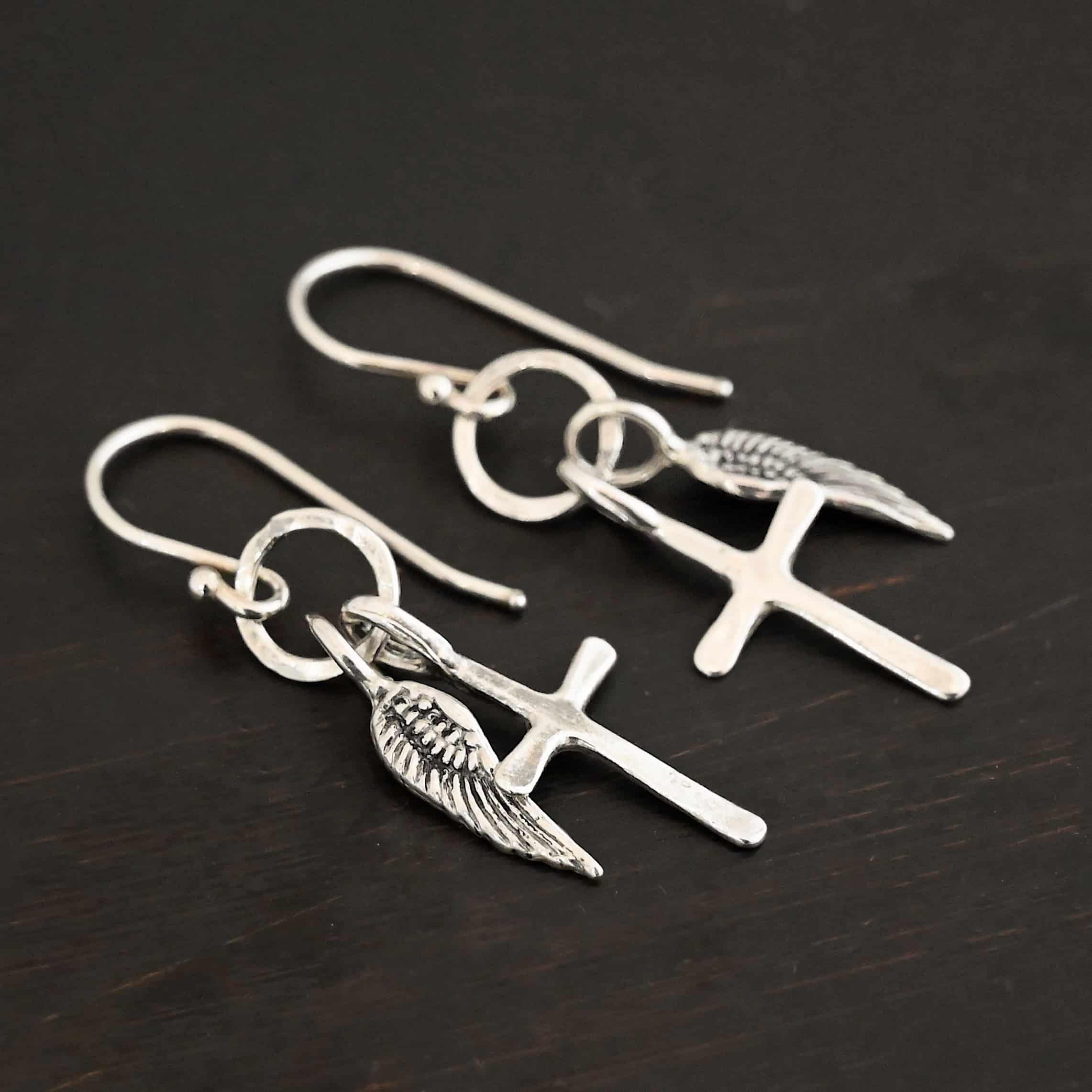 Strength Earrings Photo of the "Strength" Earrings. Made out of sterling silver. There are a cross and angel wing charm on each earring. Both charms are small, and attached to a small hammered sterling silver loop that the French hook ear wires are attached to on the top. Earrings are shiny and laying against a dark background.