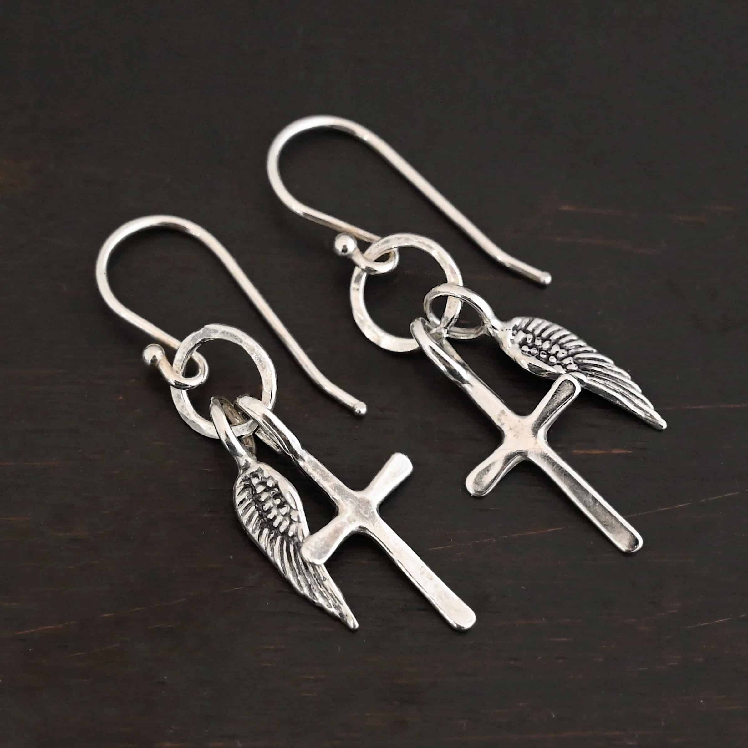 Strength Earrings Photo of the "Strength" Earrings. Made out of sterling silver. There are a cross and angel wing charm on each earring. Both charms are small, and attached to a small hammered sterling silver loop that the French hook ear wires are attached to on the top. Earrings are shiny and laying against a dark background.