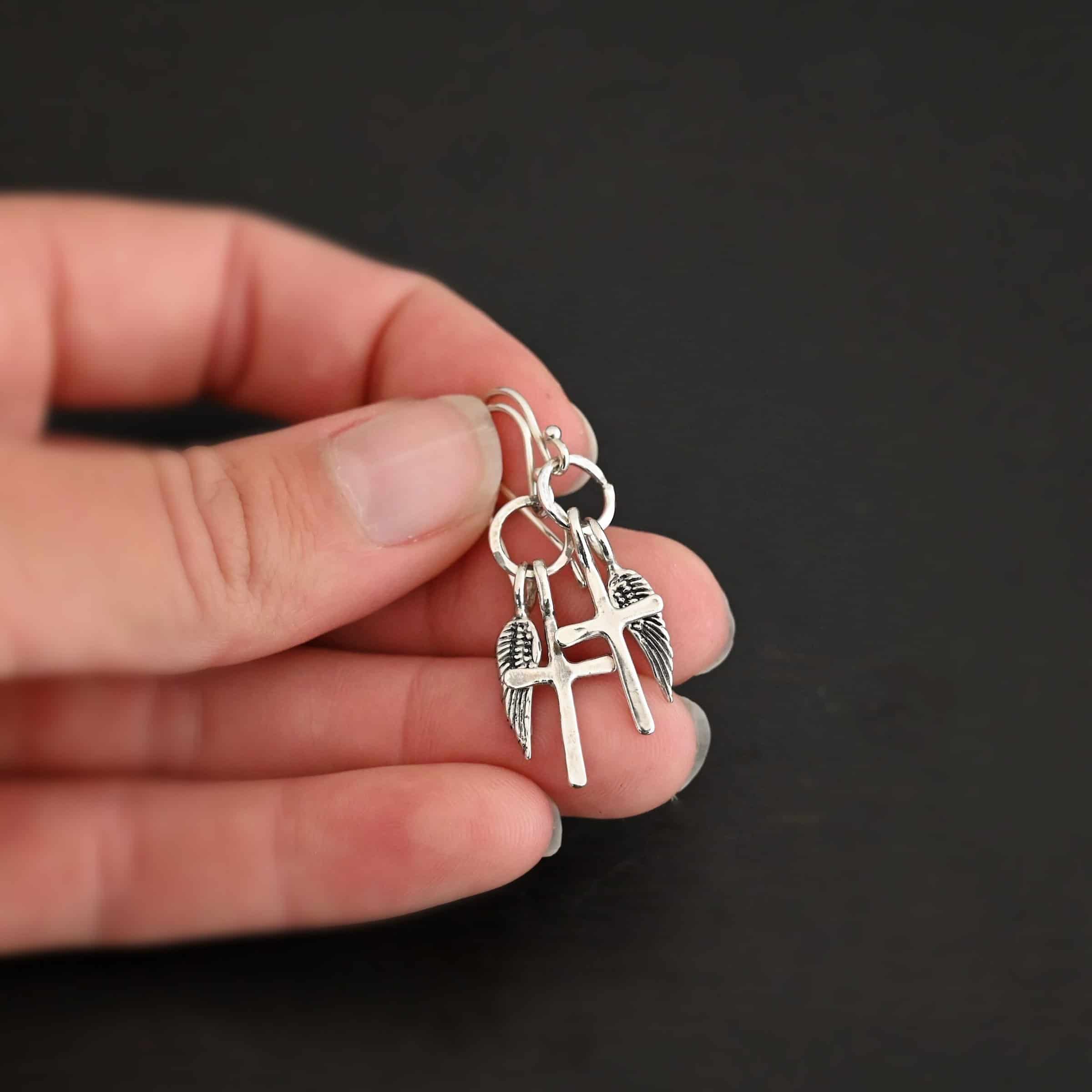 Strength Earrings Photo of the "Strength" Earrings. Made out of sterling silver. There are a cross and angel wing charm on each earring. Both charms are small, and attached to a small hammered sterling silver loop that the French hook ear wires are attached to on the top. Earrings are shiny and held in the fingertips of a hand to demonstrate size.