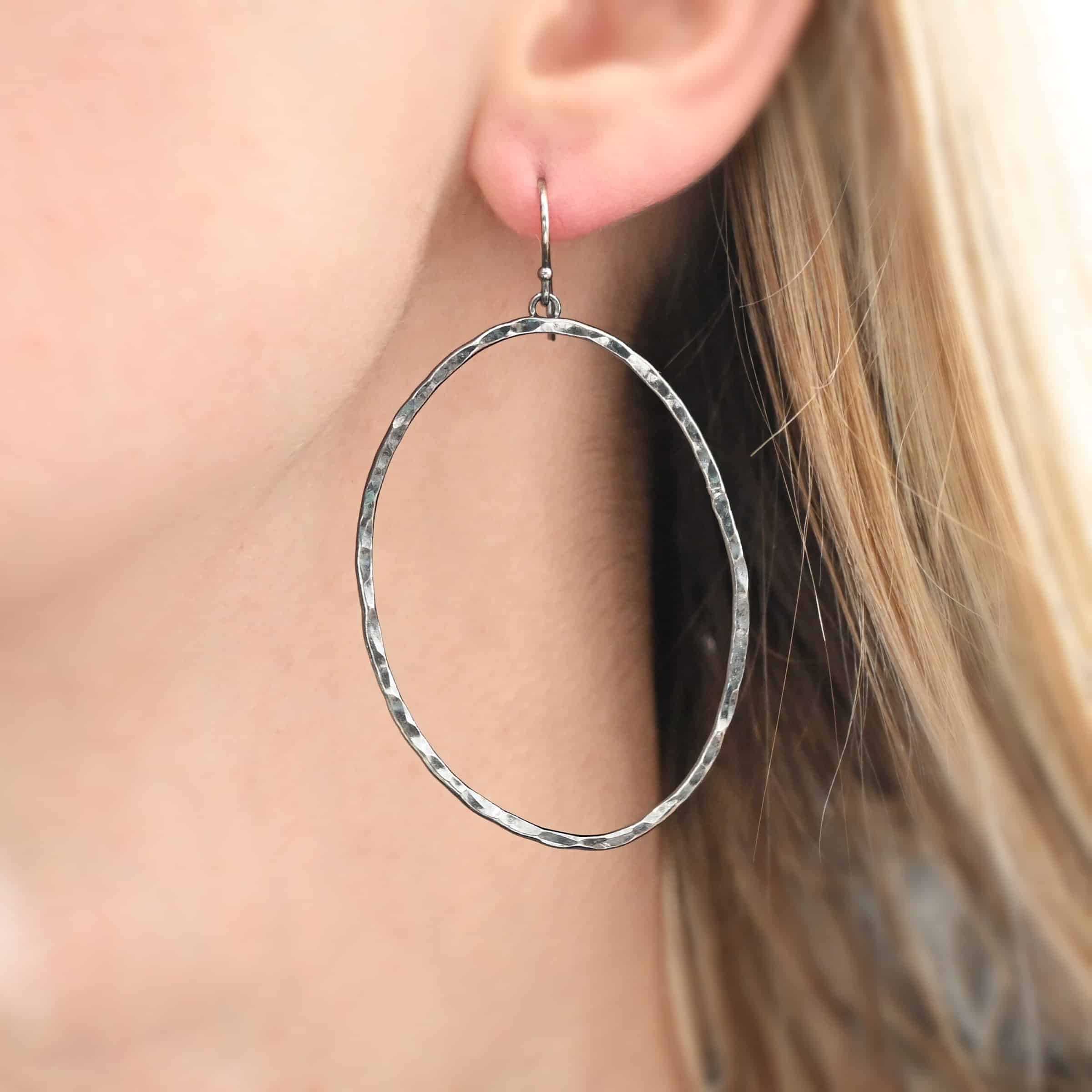 Photo of the "Haines Beach" Hoop Earring as shown on a model. Made out of sterling silver and oxidized. Earring has a french hook ear wire. Attached to the bottom of the ear wire is a small loop. The small loop is soldered to the back top of large hoop earrings that are hammered and oxidized ovals.