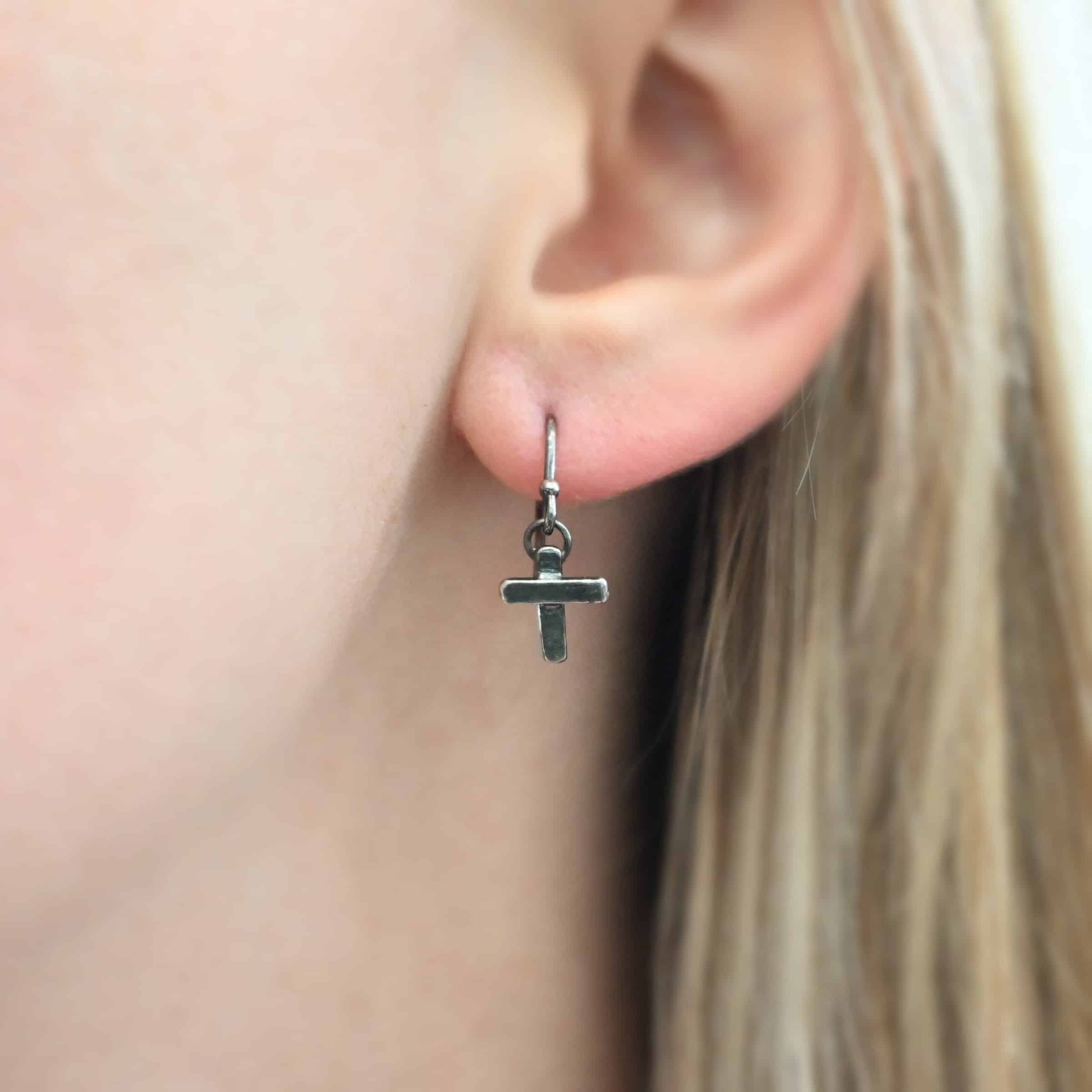 Photo of the "Old Rugged Cross" Earring as shown on a model. Made out of sterling silver and oxidized. Earring has a french hook ear wire with a jump ring on the bottom of it. Soldered to the jump ring is a small handmade cross charm. The cross has a small rectangle piece on the back, and a shorter rectangle piece soldered on top to the back piece. The shorter piece goes across the center of the back piece.