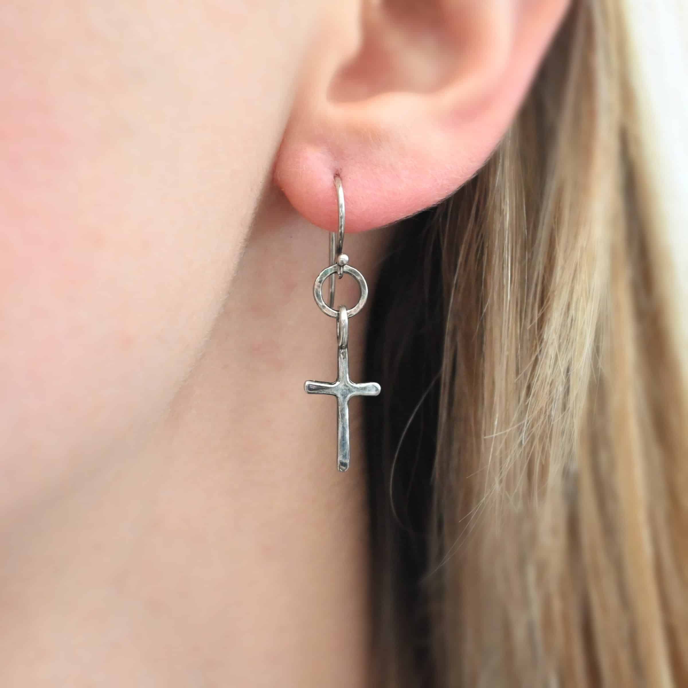 Photo of the "Redeemer" Earring as shown on a model. Made out of sterling silver and shiny. Earring has a french hook ear wire. There are medium sized, hammered circles attached to the bottom of the ear wire. Attached to the circles on the bottom is a cross charm with a soldered loop on the top that goes through the medium size circle.