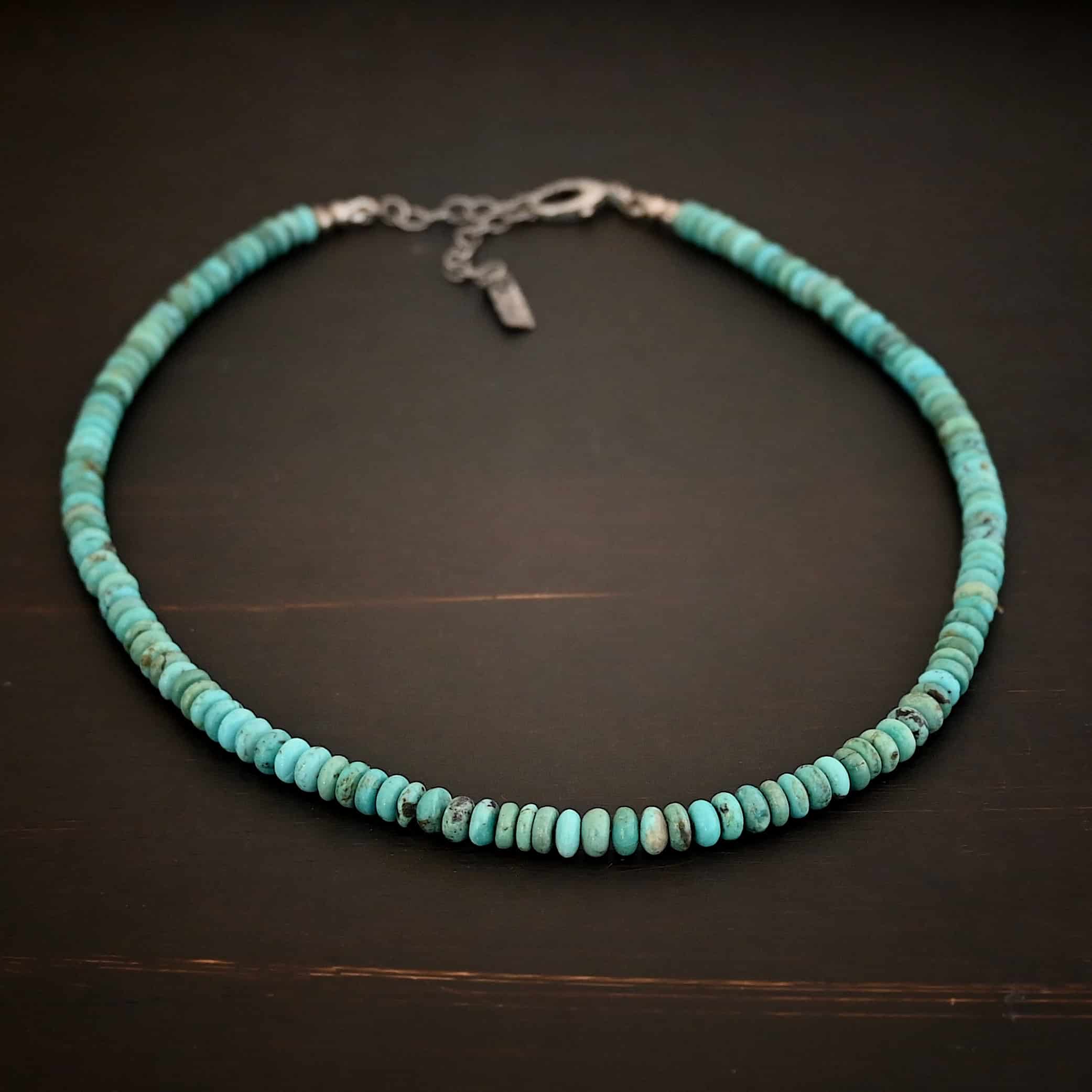 Photo of the "Hadley" Necklace. Made out of sterling silver and turquoise. The entire necklace has small, oval shaped turquoise beads strung around it. The ends of each side have two silver beads with crimps to keep the turquoise tight together. Necklace has a lobster claw clasp with a sterling silver extension. Necklace has a small rectangle tag on the end of it that has "Wild Prairie Silver" stamped into it. Necklace is lying against a dark background.