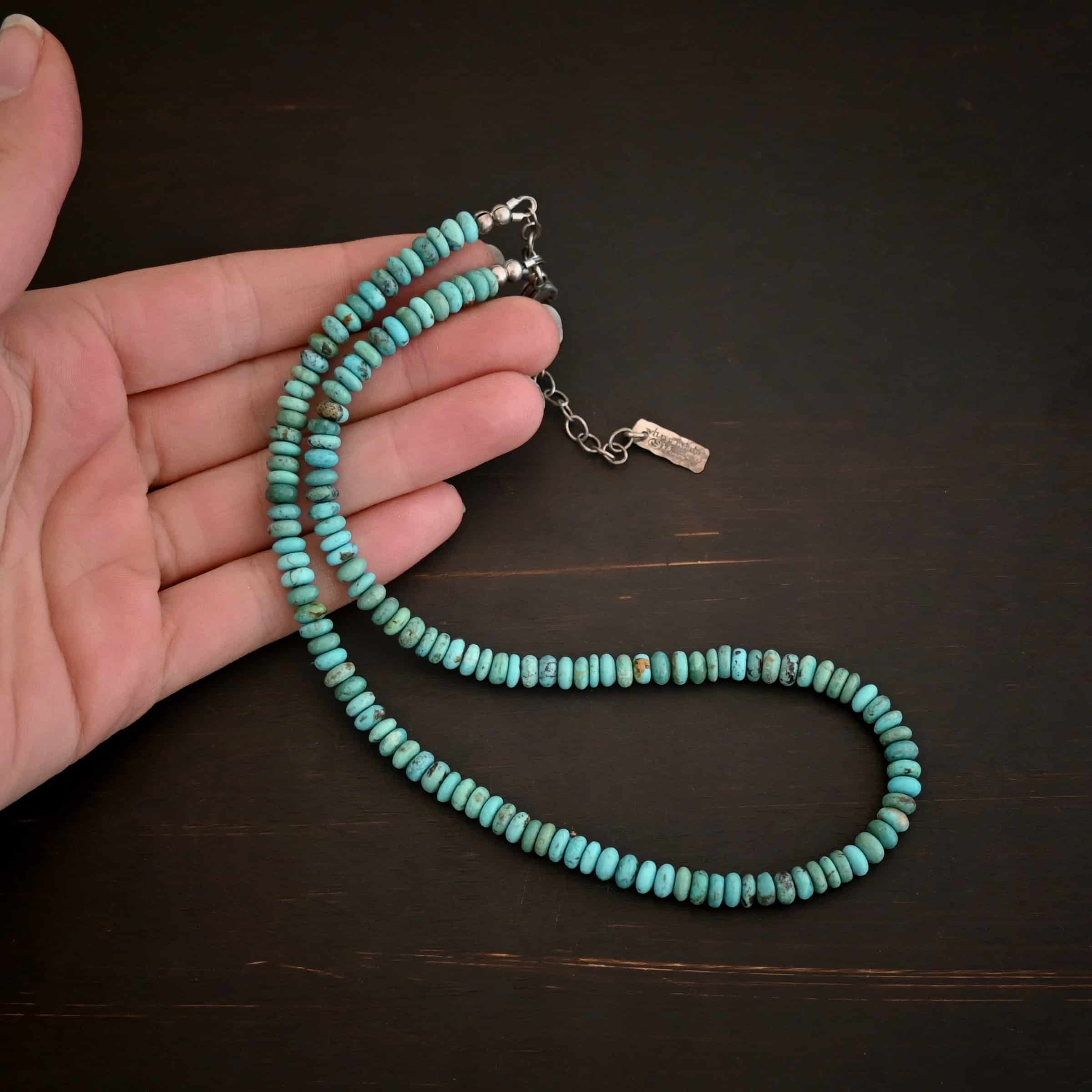 Photo of the "Hadley" Necklace. Made out of sterling silver and turquoise. The entire necklace has small, oval shaped turquoise beads strung around it. The ends of each side have two silver beads with crimps to keep the turquoise tight together. Necklace has a lobster claw clasp with a sterling silver extension. Necklace has a small rectangle tag on the end of it that has "Wild Prairie Silver" stamped into it. Necklace is lying against the fingertips of a hand on a dark background.