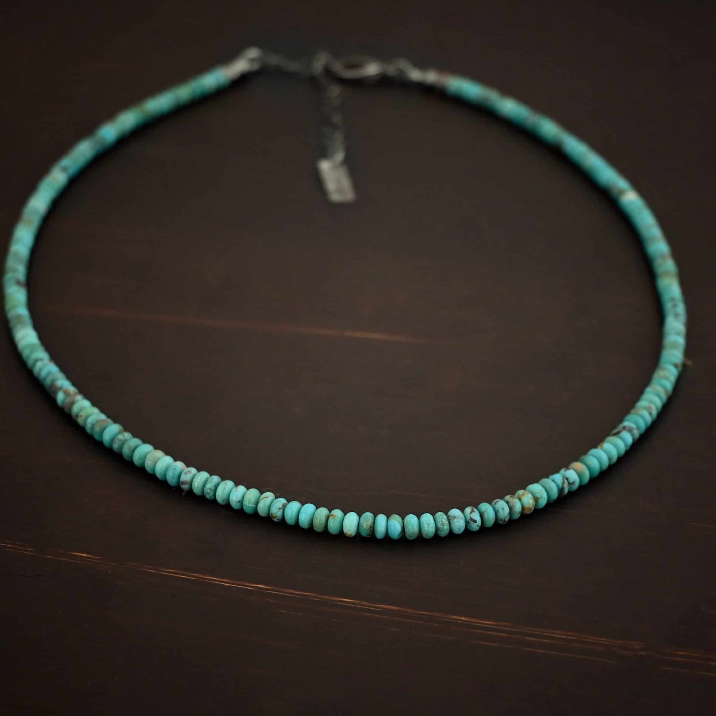 Photo of the "Hadley" Necklace. Made out of sterling silver and turquoise. The entire necklace has small, oval shaped turquoise beads strung around it. The ends of each side have two silver beads with crimps to keep the turquoise tight together. Necklace has a lobster claw clasp with a sterling silver extension. Necklace has a small rectangle tag on the end of it that has "Wild Prairie Silver" stamped into it. Necklace is lying against a dark background.