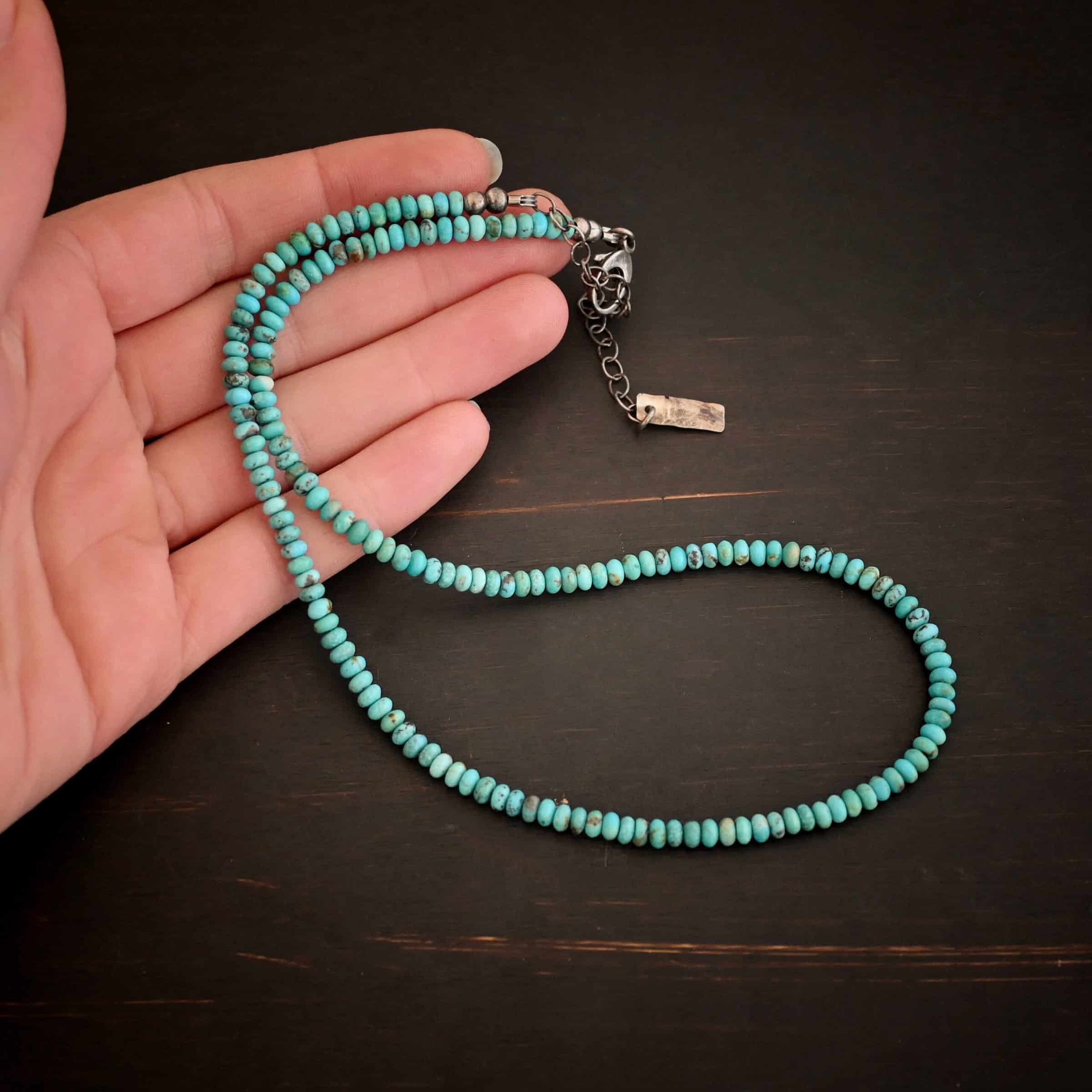 Photo of the "Hadley" Necklace. Made out of sterling silver and turquoise. The entire necklace has small, oval shaped turquoise beads strung around it. The ends of each side have two silver beads with crimps to keep the turquoise tight together. Necklace has a lobster claw clasp with a sterling silver extension. Necklace has a small rectangle tag on the end of it that has "Wild Prairie Silver" stamped into it. Necklace is lying against the fingertips of a hand to demonstrate size.