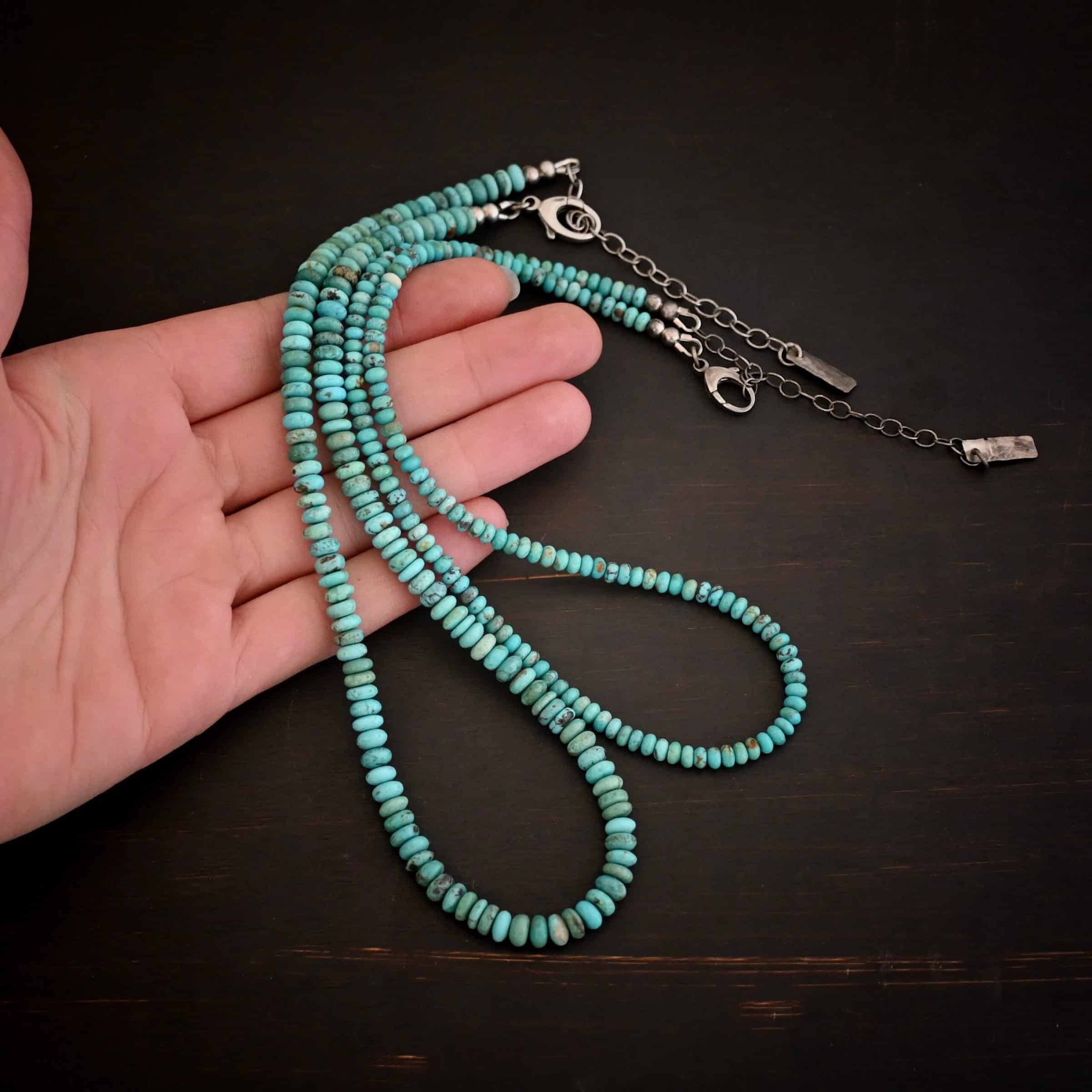 Photo of the "Hadley" Necklace with different sized beads. Made out of sterling silver and turquoise. The entire necklace has small, oval shaped turquoise beads strung around it. The ends of each side have two silver beads with crimps to keep the turquoise tight together. Necklace has a lobster claw clasp with a sterling silver extension. Necklace has a small rectangle tag on the end of it that has "Wild Prairie Silver" stamped into it. The larger beads are on the left. Necklaces are lying against the fingertips of a hand to demonstrate size.