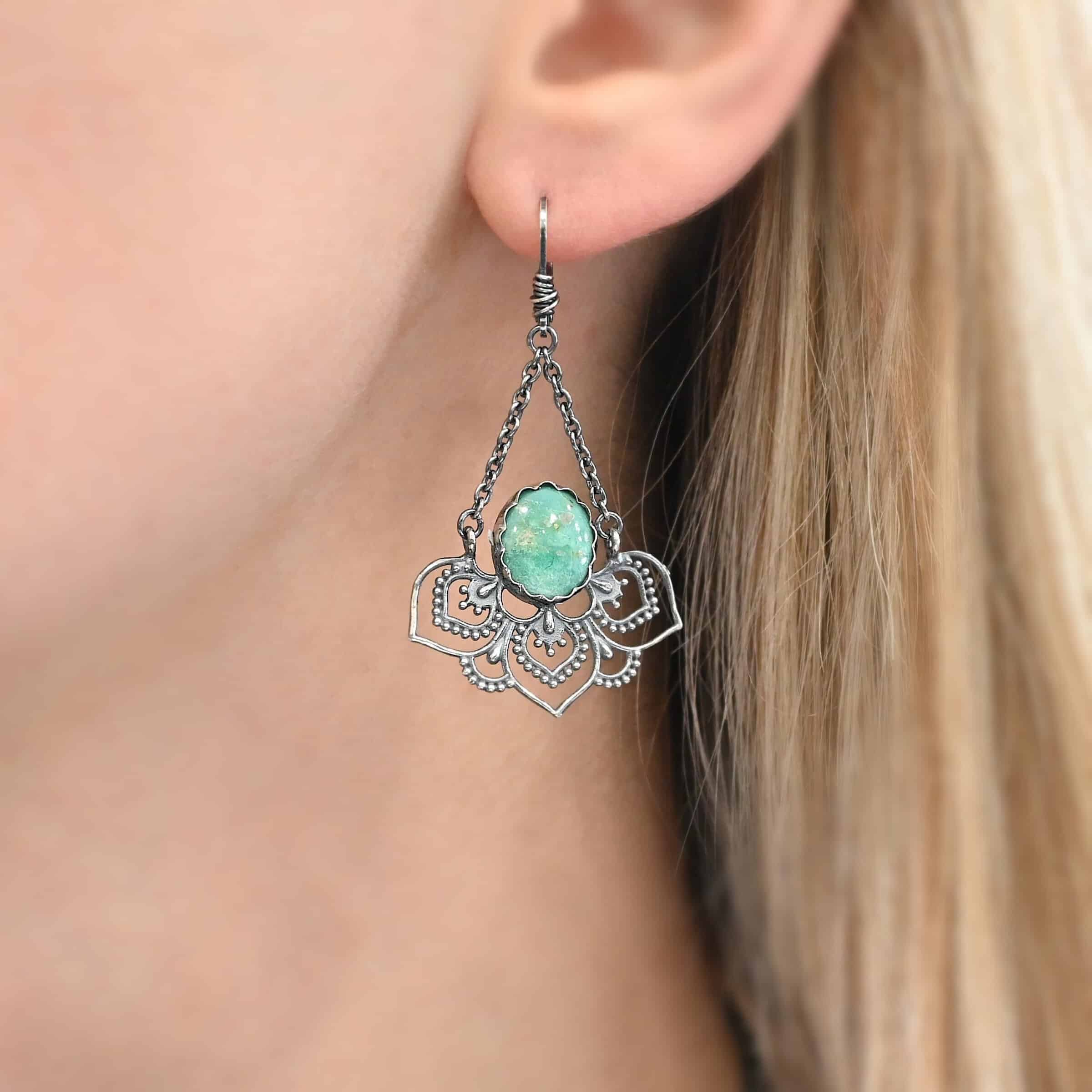 Photo of the "Hotel Congress" Earring as shown on a model. Made out of sterling silver and turquoise. Earring has a leverback ear wire with a jump ring attached to the bottom of it. There are two small strands of silver that are attached to the jump ring. The strands go opposite ways to create a triangle shape. Attached to each end of the strands of silver is a half flower shaped pendant. The pendant is made out of sterling silver, and has three large leaves and two semicircles in between the three leaves. There is an oval turquoise that is attached on the bottom to all three leaves, and the turquoise sits in the center of all three of them.