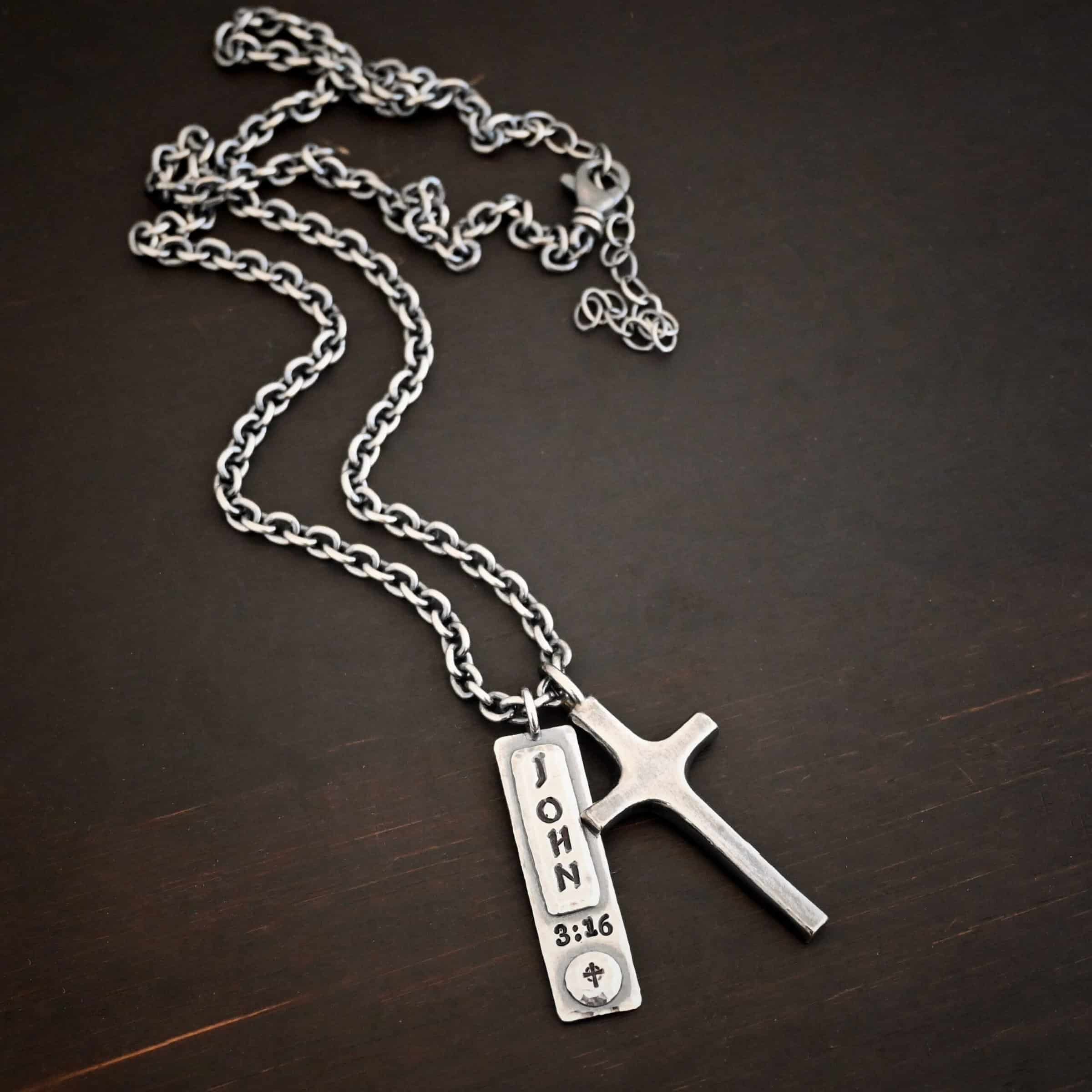 John 3:16 Necklace Photo of the "John 3:16" Necklace. Made out of sterling silver. There is a medium sized cross that is made out of smooth, thick sterling silver. It is oxidized and brushed lightly and has a loop soldered to the top of it. There is a rectangle tag next to it with the word "John" stamped into it horizontally and "3:16" beneath it stamped into the necklace vertically. There is a small cross stamped into the bottom of the necklace on a circle of sterling silver. There is a chain that goes through the top of the necklace. Necklace is lying against a dark background.