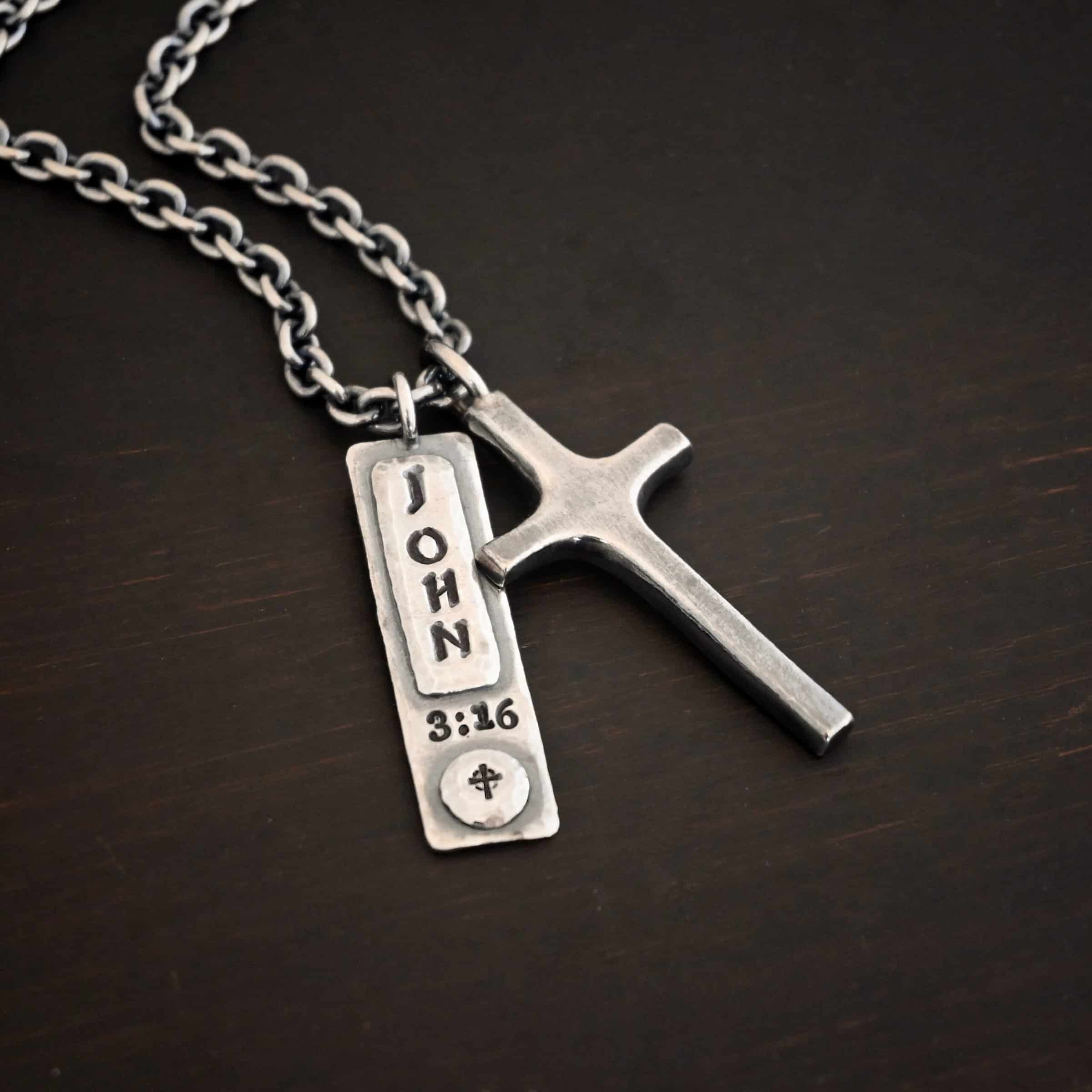John 3:16 Necklace Photo of the "John 3:16" Necklace. Made out of sterling silver. There is a medium sized cross that is made out of smooth, thick sterling silver. It is oxidized and brushed lightly and has a loop soldered to the top of it. There is a rectangle tag next to it with the word "John" stamped into it horizontally and "3:16" beneath it stamped into the necklace vertically. There is a small cross stamped into the bottom of the necklace on a circle of sterling silver. There is a chain that goes through the top of the necklace. Necklace is lying against a dark background.