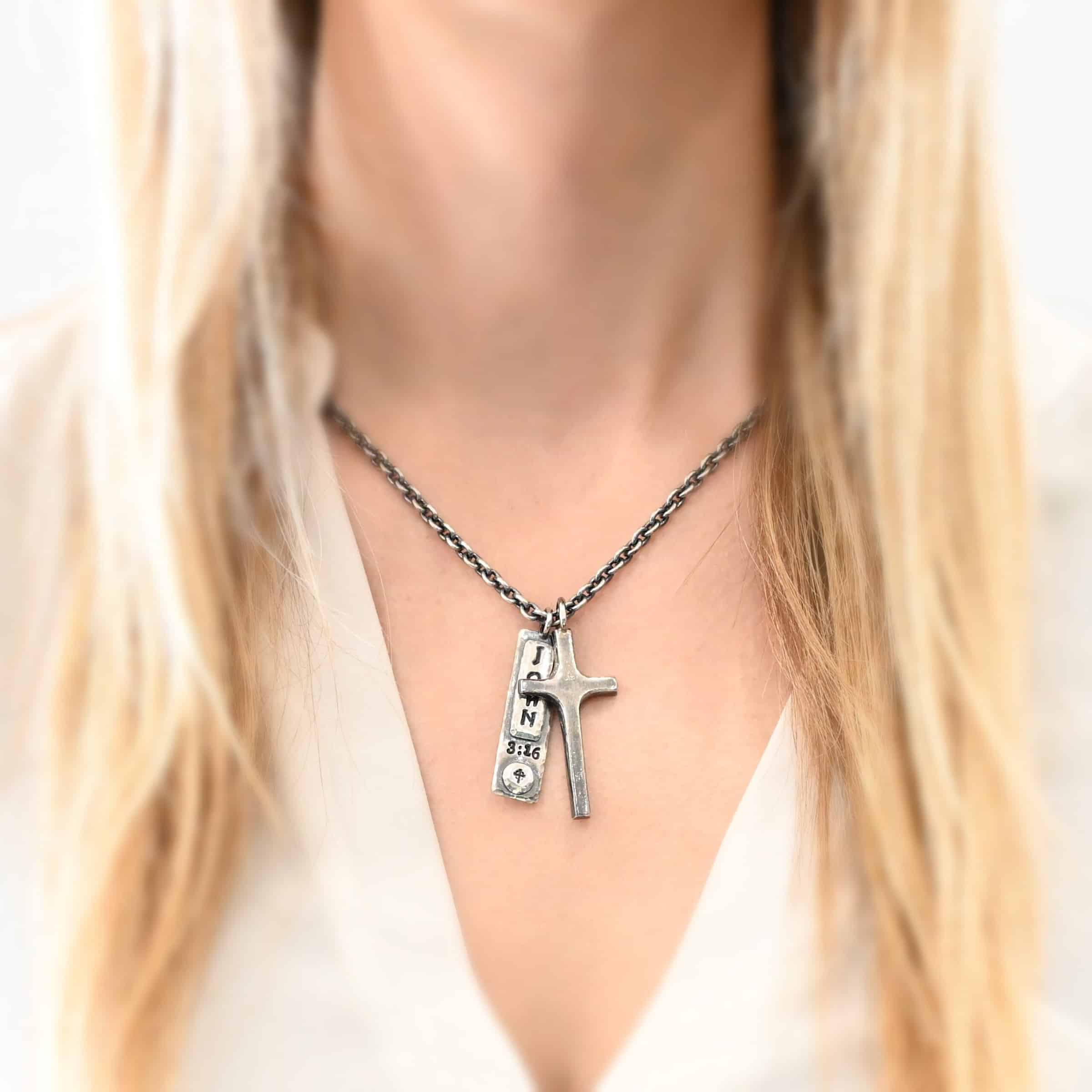 John 3:16 Necklace Photo of the "John 3:16" Necklace. Made out of sterling silver. There is a medium sized cross that is made out of smooth, thick sterling silver. It is oxidized and brushed lightly and has a loop soldered to the top of it. There is a rectangle tag next to it with the word "John" stamped into it horizontally and "3:16" beneath it stamped into the necklace vertically. There is a small cross stamped into the bottom of the necklace on a circle of sterling silver. There is a chain that goes through the top of the necklace. Necklace is worn by a model to demonstrate size.
