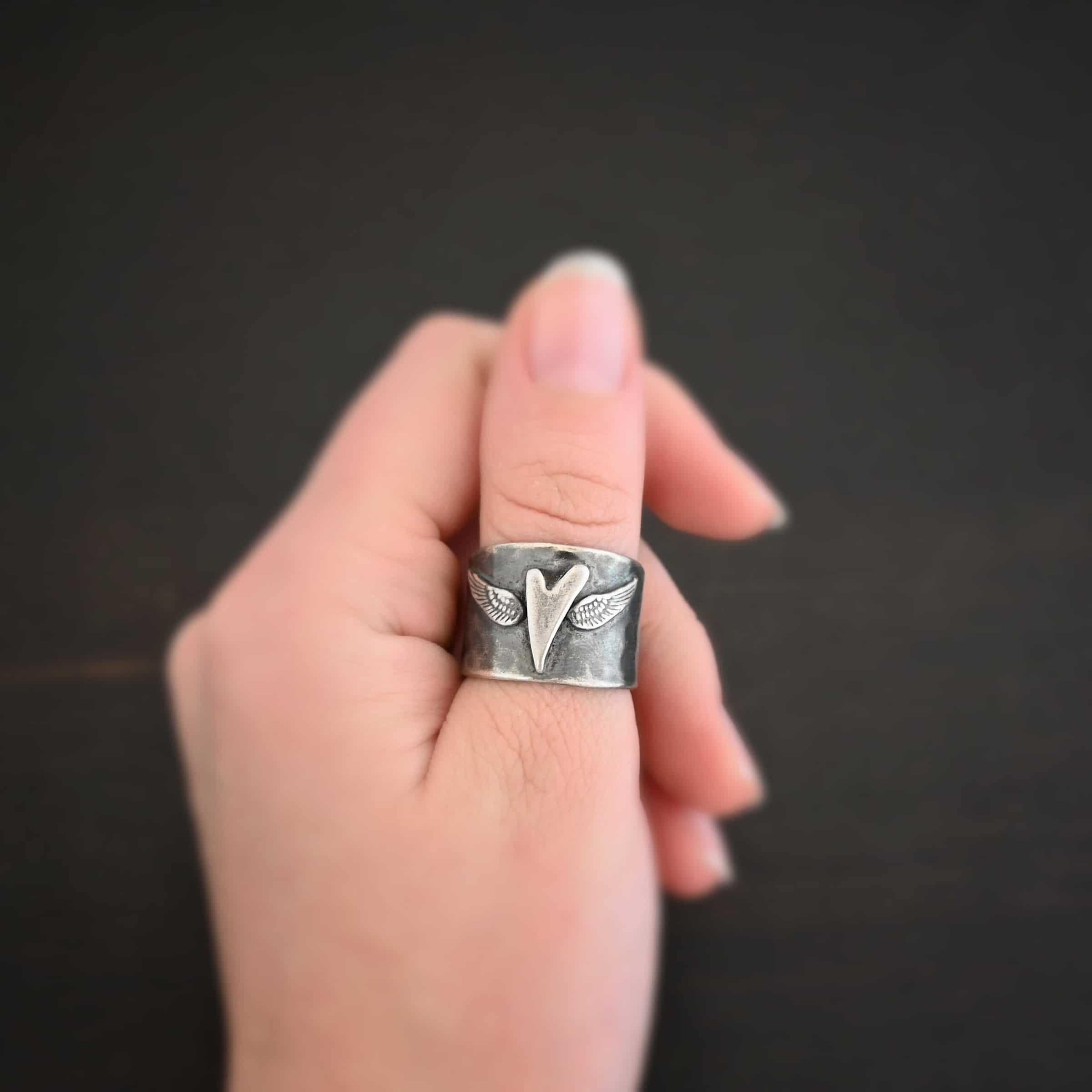 Close up of the "Wild Prairie" Ring. Made out of sterling silver. There is a silver heart in the center of the ring. It is a tall heart that stretches from almost the top of the ring to the bottom. There are sterling silver angel wings sprouting from each side of the heart. Ring is worn by a model to demonstrate size.