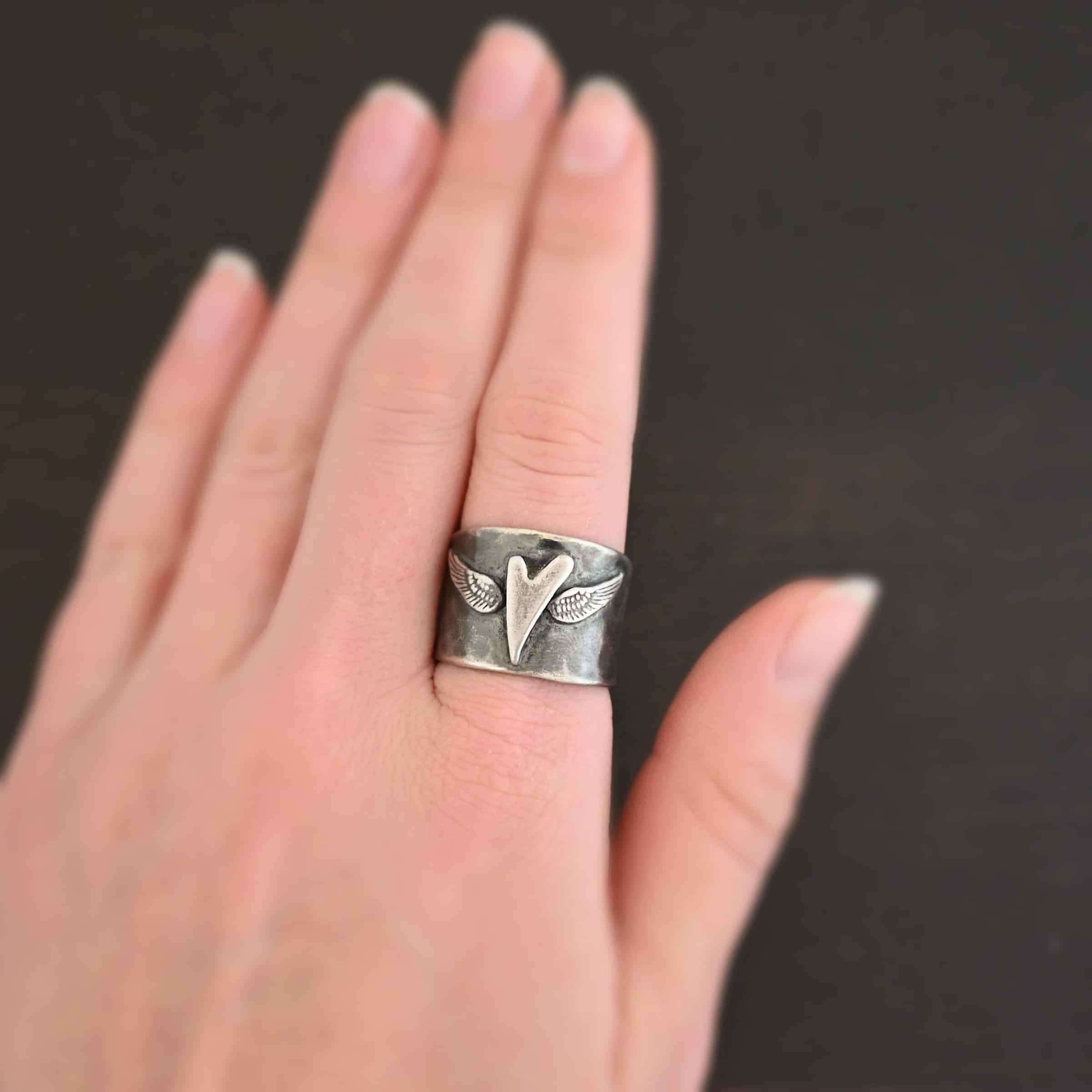 Close up of the "Wild Prairie" Ring. Made out of sterling silver. There is a silver heart in the center of the ring. It is a tall heart that stretches from almost the top of the ring to the bottom. There are sterling silver angel wings sprouting from each side of the heart. Ring is worn by a model to demonstrate size.