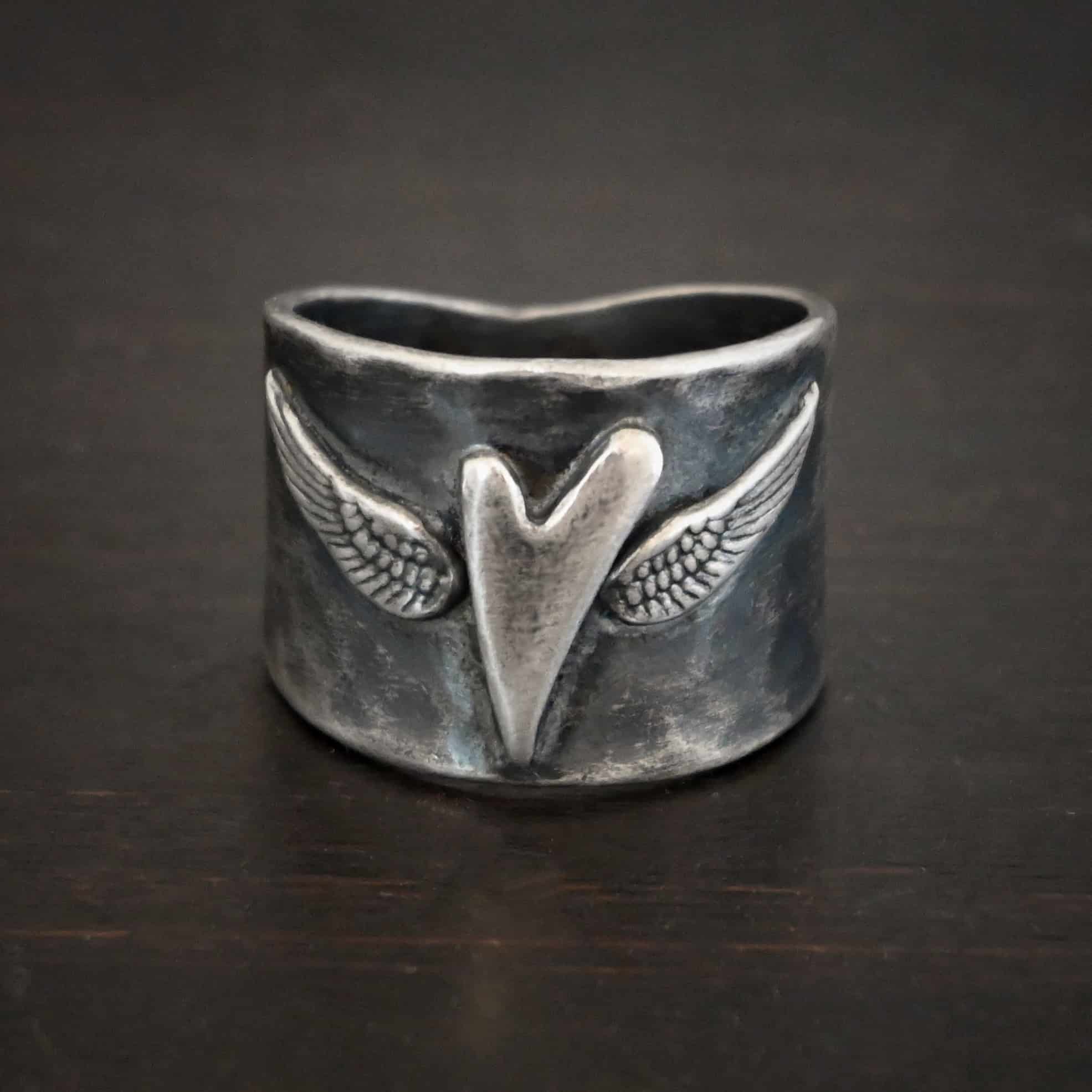 Close up of the "Wild Prairie" Ring. Made out of sterling silver. There is a silver heart in the center of the ring. It is a tall heart that stretches from almost the top of the ring to the bottom. There are sterling silver angel wings sprouting from each side of the heart. Ring is sitting against a dark background.