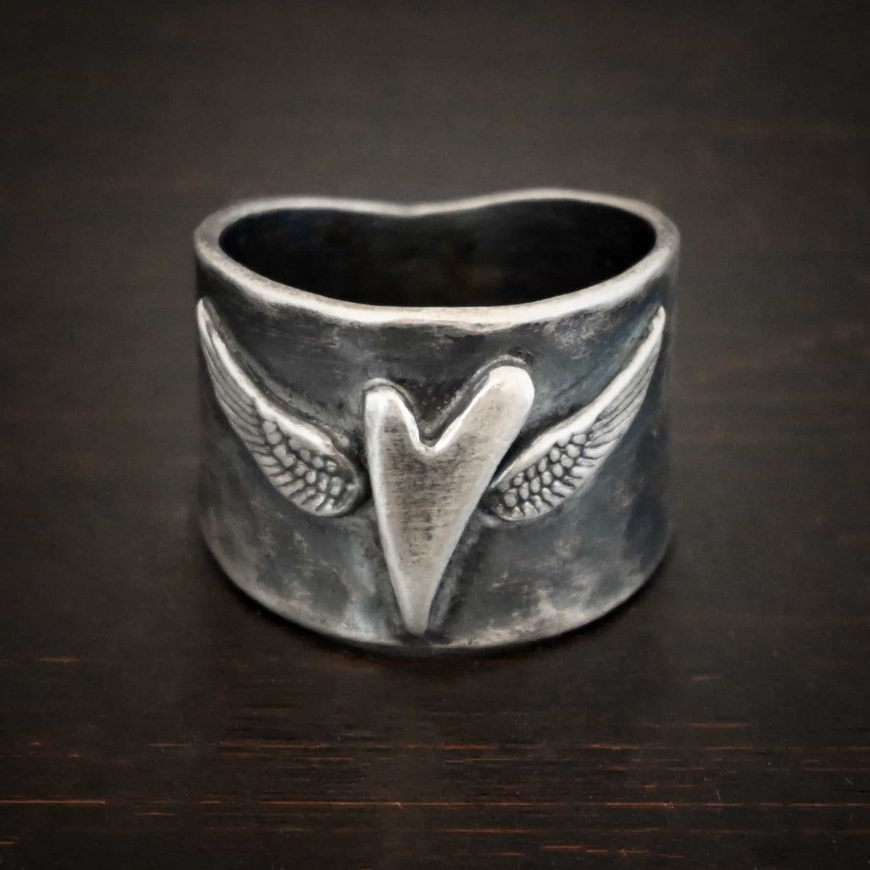 Close up of the "Wild Prairie" Ring. Made out of sterling silver. There is a silver heart in the center of the ring. It is a tall heart that stretches from almost the top of the ring to the bottom. There are sterling silver angel wings sprouting from each side of the heart. Ring is sitting against a dark background.