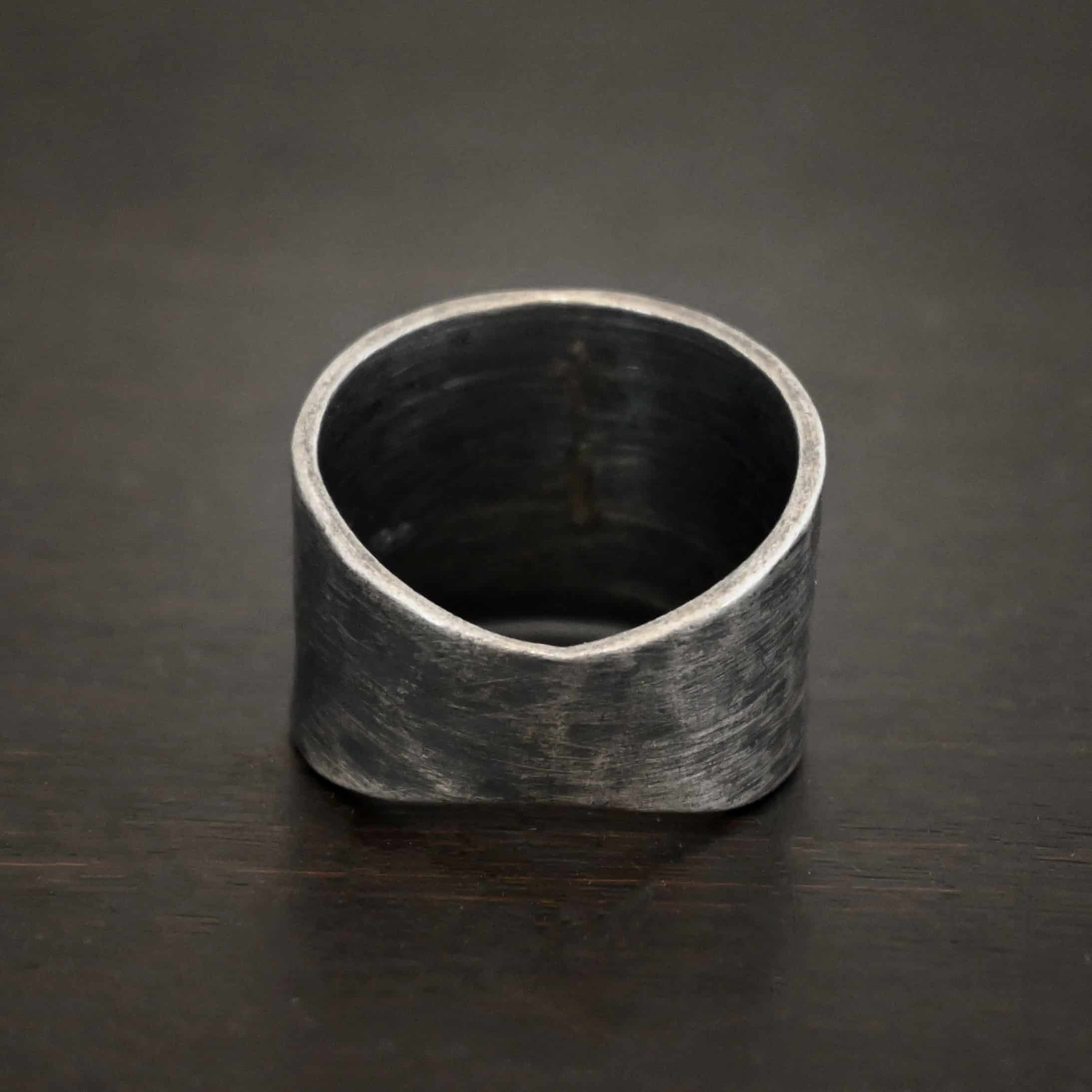 Close up of the back of the "Wild Prairie" Ring. Made out of sterling silver. The back of the ring is smooth, and tapers, so it is smaller than the front of the ring. Ring is oxidized and sitting against a dark background.